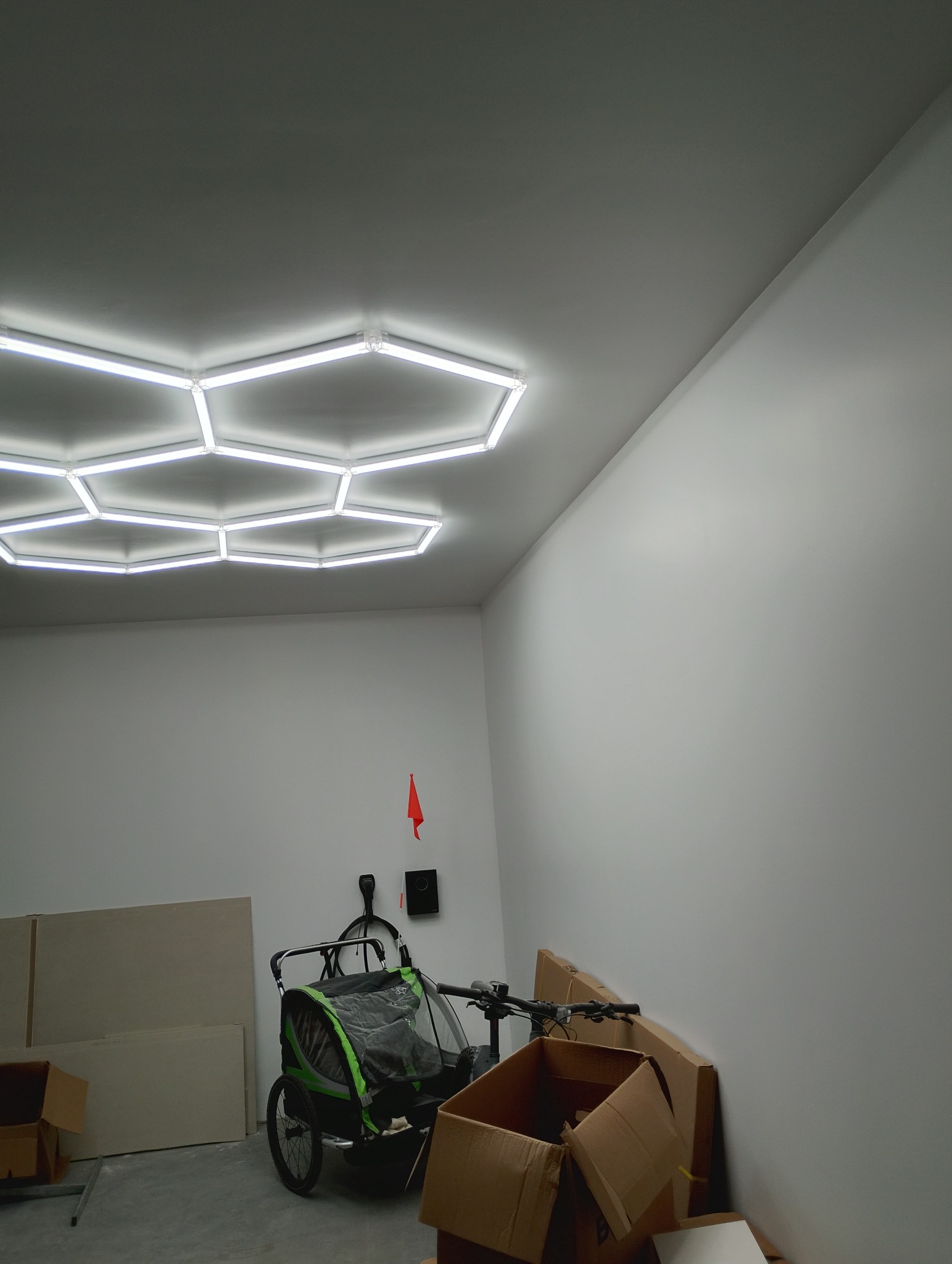 Białe ściany garażu z zamontowanym oświetleniem sufitowym LED w kształcie plastra miodu. Na podłodze kartony, przyczepka rowerowa i płyty gipsowe.