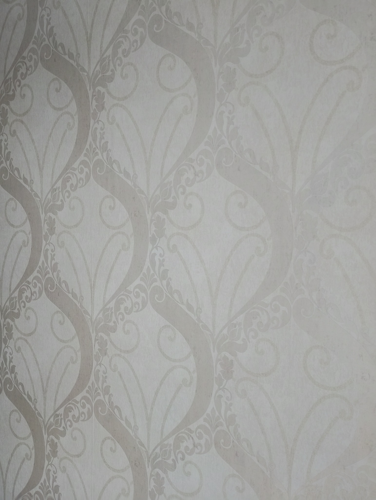 Delikatna, beżowo-biała tapeta w ornamenty roślinne i geometryczne, tworząca elegancki wzór na ścianie. Subtelne przejścia tonalne podkreślają fakturę.