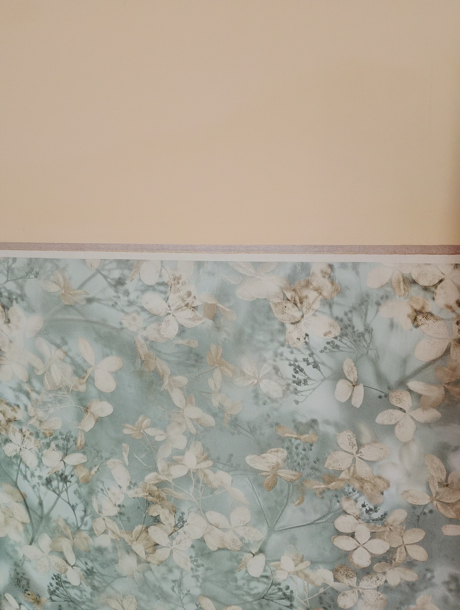 Ściana z tapetą w kwiaty hortensji, oddzielona od beżowego sufitu szarą listwą. Delikatny wzór i pastelowe kolory tworzą spokojną atmosferę.