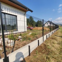 MT OGRODZENIA Maciej Tokarczyk - Nowoczesne ogrodzenie panelowe w kolorze czarnym, osadzone na betonowej podmurówce. W tle dom z dużym oknem i błękitne niebo.