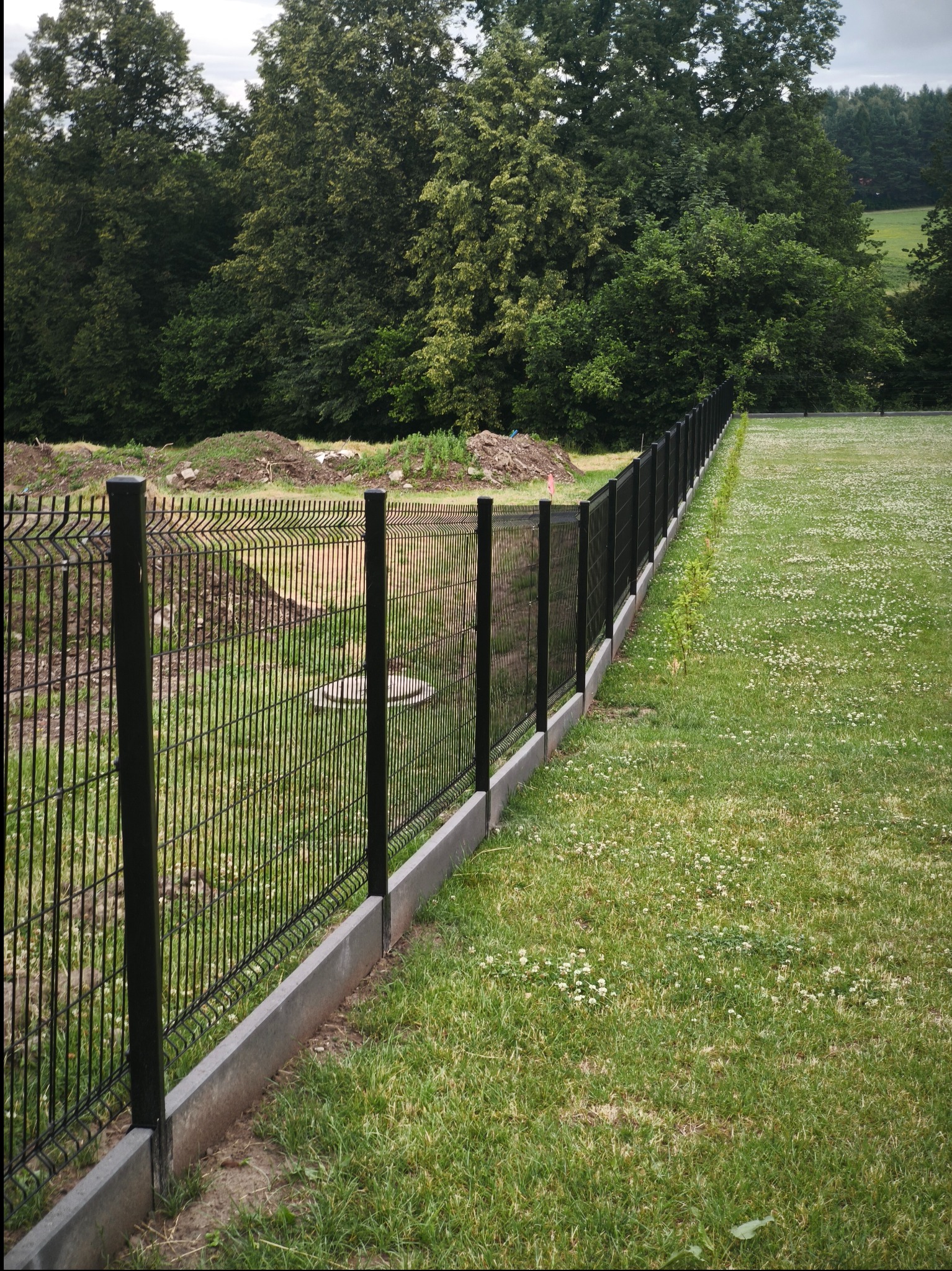 Nowoczesne ogrodzenie panelowe z podmurówką betonową, czarne słupki, widok na zielony trawnik i drzewa w tle, świeżo zamontowane, widoczne ślady prac ziemnych.