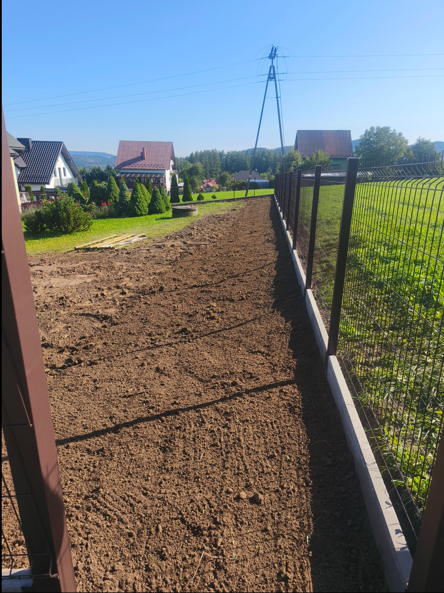 Nowoczesne ogrodzenie panelowe z podmurówką w Przysietnicy, widok z perspektywy ziemi, z brązowymi słupkami i siatką na tle zielonej trawy i zabudowań.