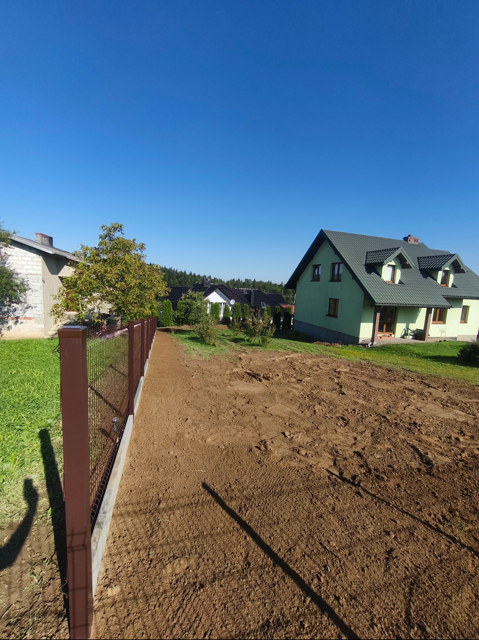Nowoczesne ogrodzenie panelowe w kolorze brązowym, z widocznymi słupkami i podmurówką, na tle zielonego trawnika i domów jednorodzinnych w słoneczny dzień.