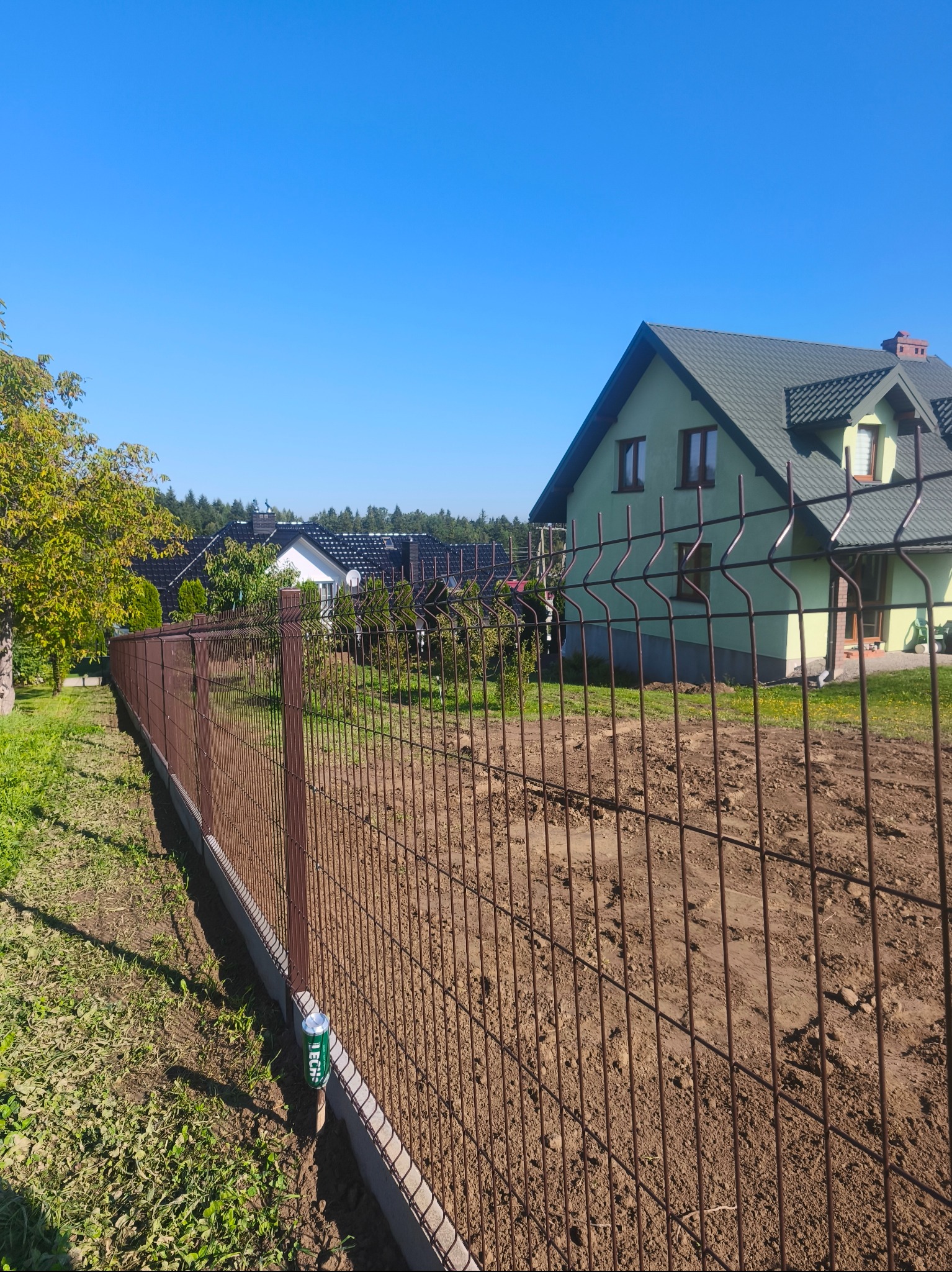 Brązowe ogrodzenie panelowe na fundamencie, z widocznym domem w tle. Na pierwszym planie puszka po napoju przy słupku.