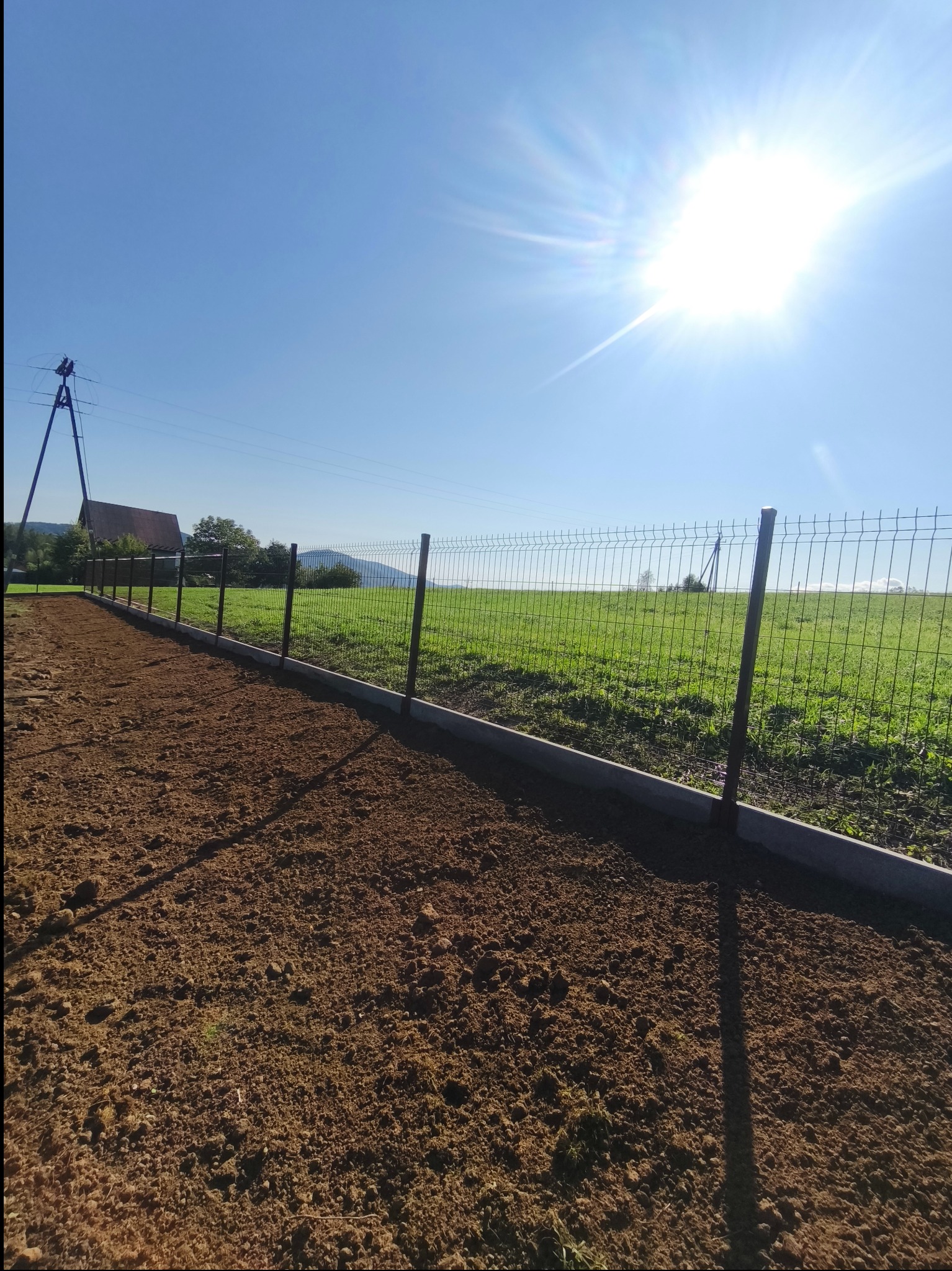 Nowoczesne ogrodzenie panelowe z podmurówką betonową na tle pola i błękitnego nieba z intensywnym słońcem. Widoczna linia energetyczna w oddali.