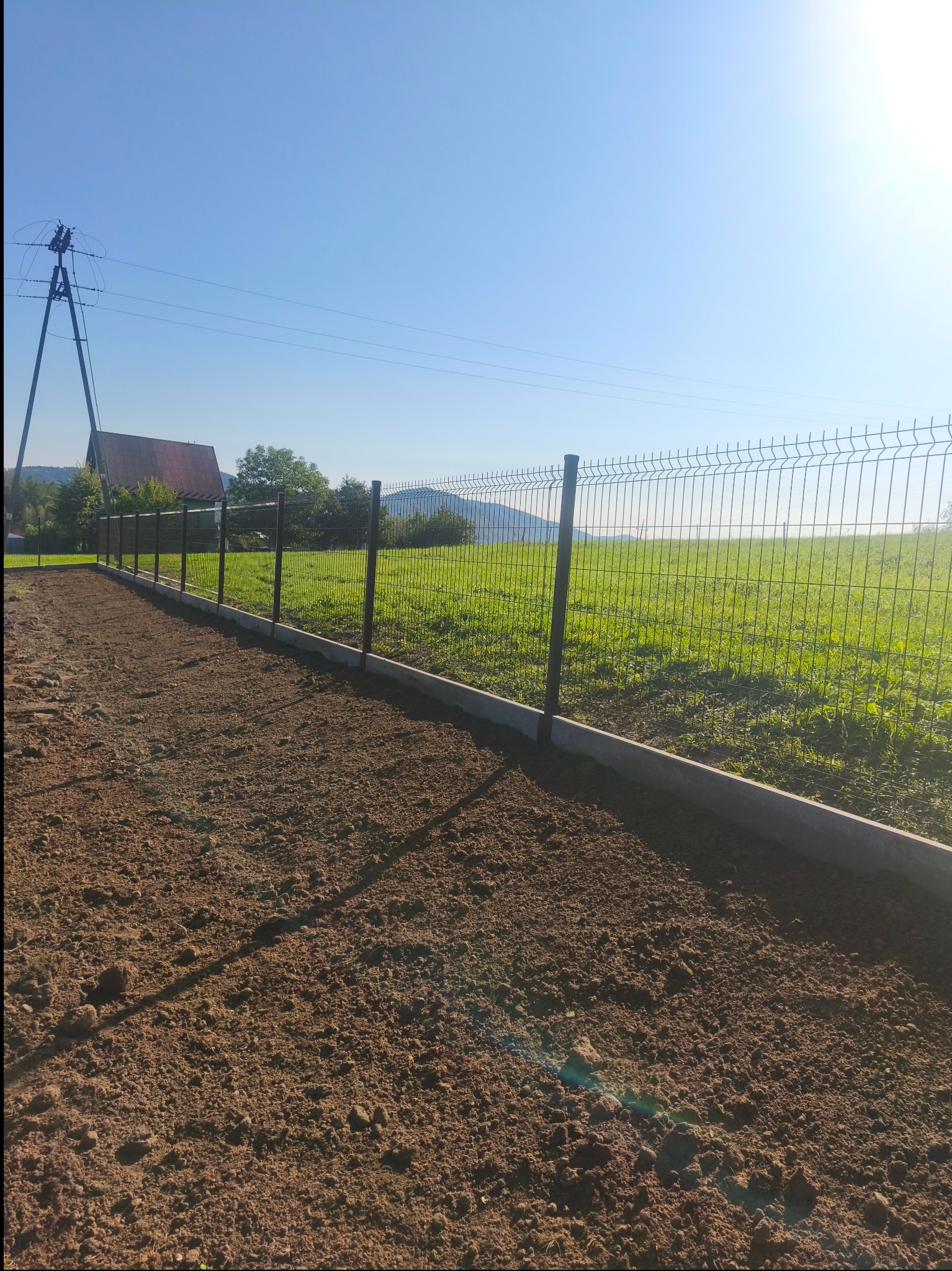 Montaż ogrodzenia panelowego na fundamencie betonowym na tle zielonego pola i błękitnego nieba. Widoczny słup elektryczny w oddali.