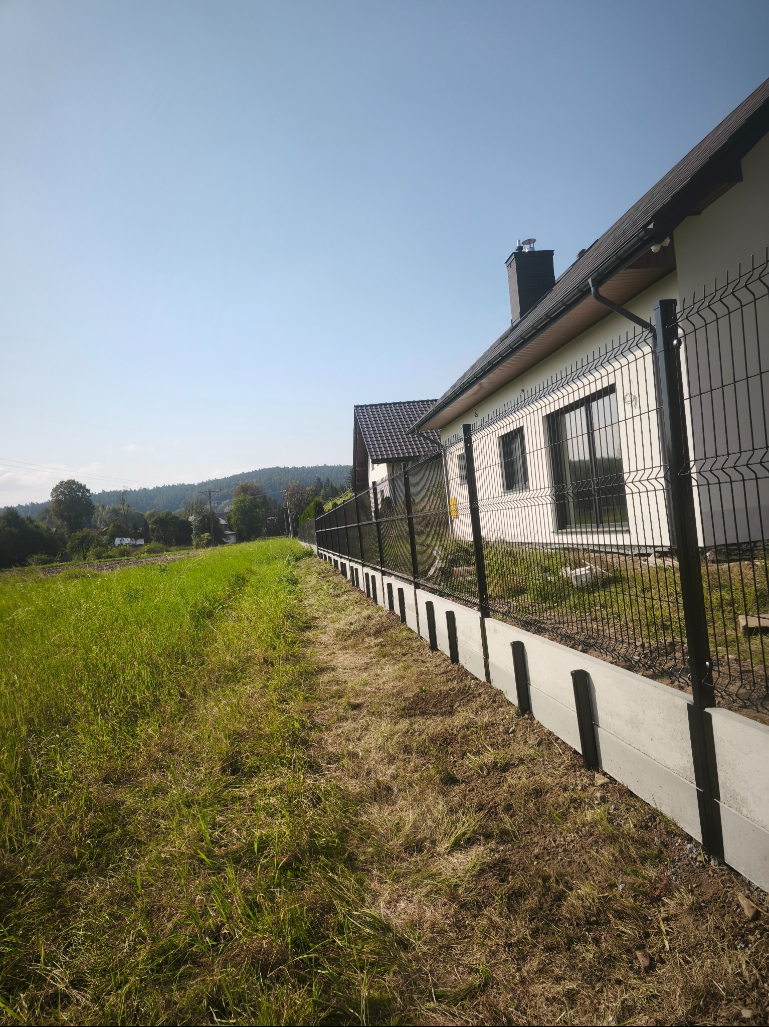 Nowoczesne ogrodzenie panelowe w kolorze czarnym, zamontowane na betonowej podmurówce, otacza posesję z domem na tle zieleni i błękitnego nieba.