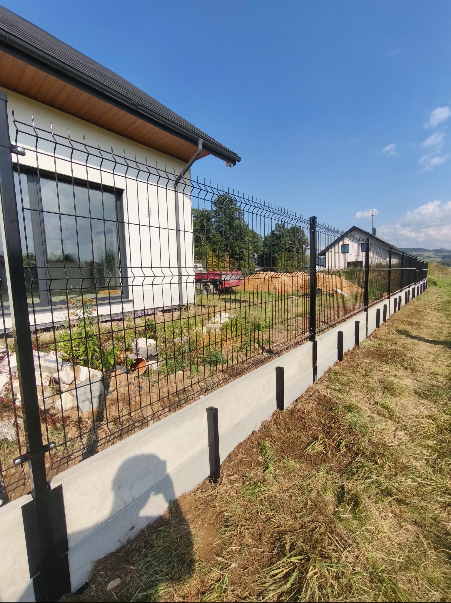 Nowoczesne ogrodzenie panelowe w kolorze czarnym, osadzone na betonowej podmurówce. W tle dom z dużym oknem i błękitne niebo.