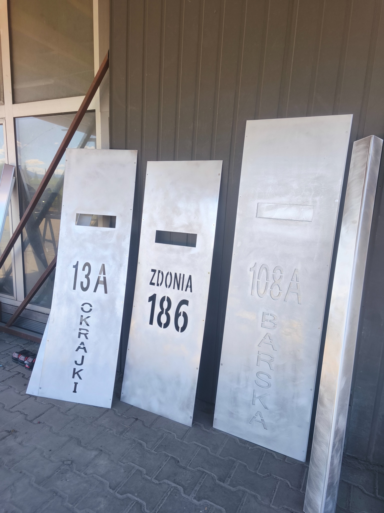 Trzy metalowe tablice adresowe w odcieniach szarości, z wyciętymi numerami i nazwami ulic: '13A Okrajki', 'Zdonią 186', '108A Barska', ustawione obok siebie.