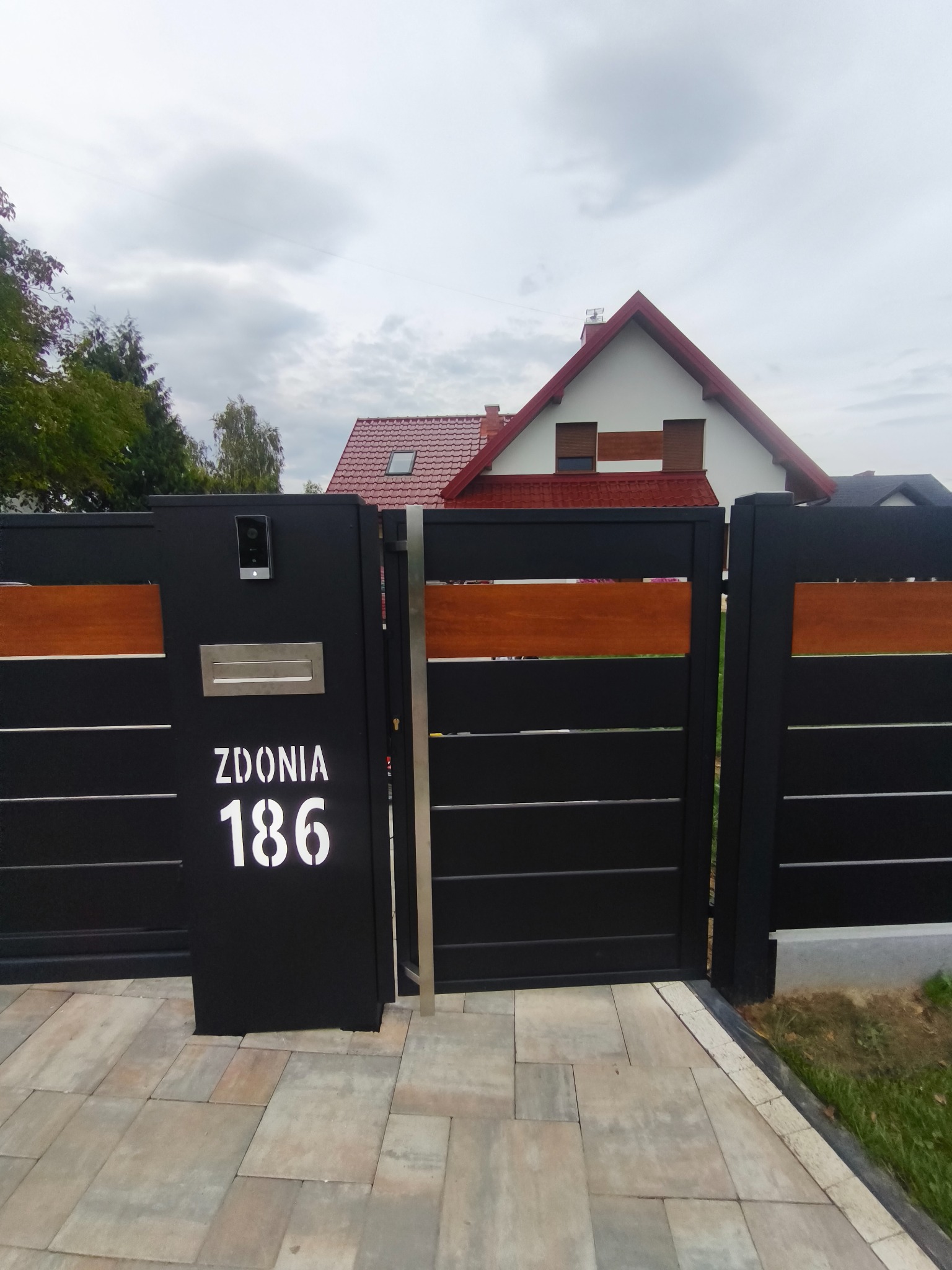 Nowoczesna brama wjazdowa z czarnymi panelami i drewnianymi akcentami. Na słupku domofon, skrzynka na listy i numer domu 'Zdonia 186'.