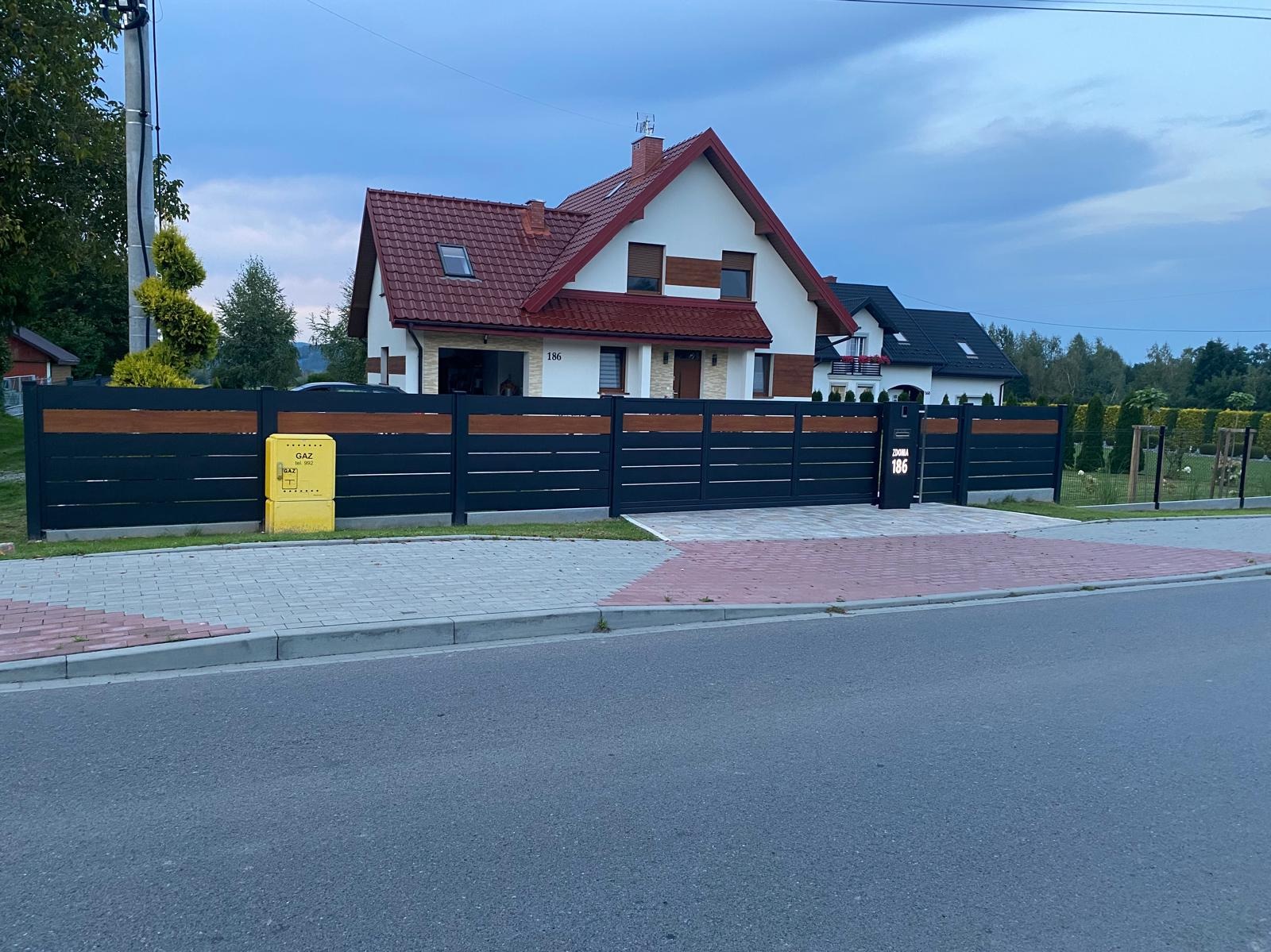 Nowoczesne ogrodzenie panelowe z drewnianymi akcentami i przesuwną bramą wjazdową, numer domu 186, skrzynka gazowa po lewej stronie.