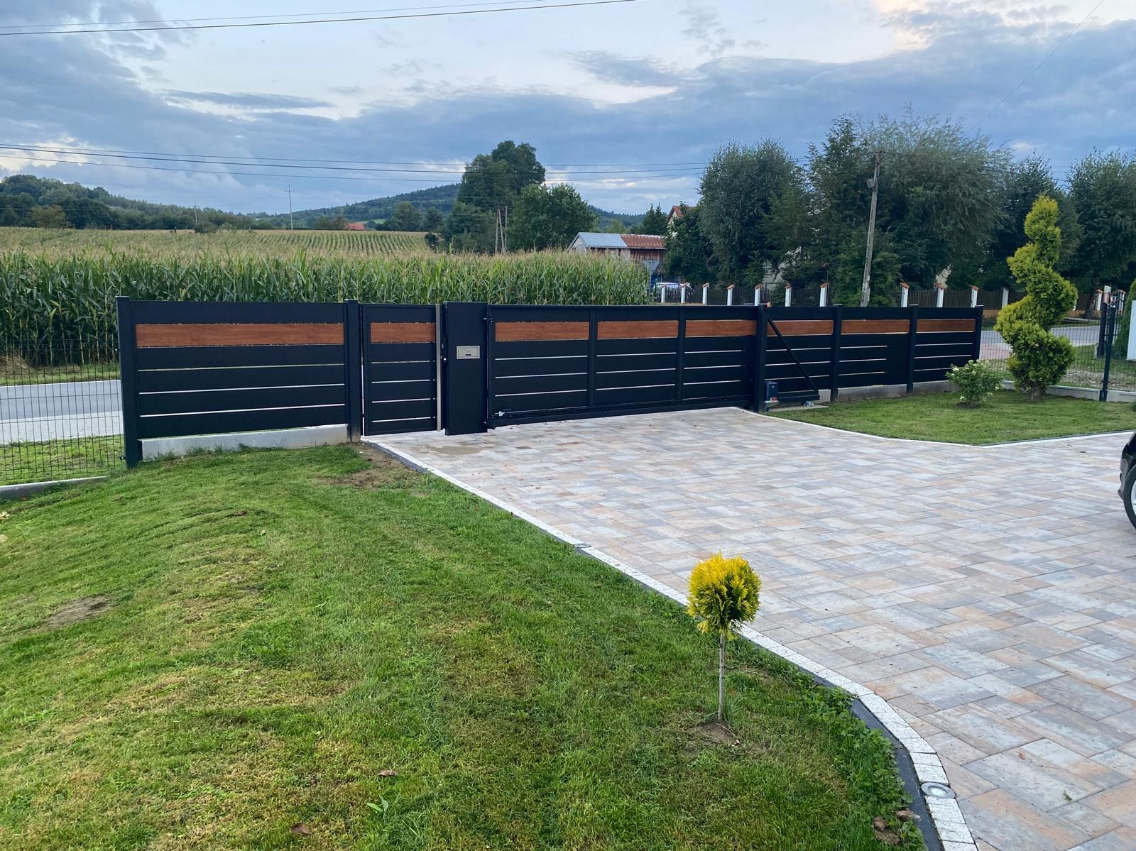 Nowoczesna, przesuwna brama wjazdowa z czarnymi panelami i drewnianymi akcentami, prowadząca na wybrukowany podjazd w otoczeniu zieleni i pola kukurydzy.