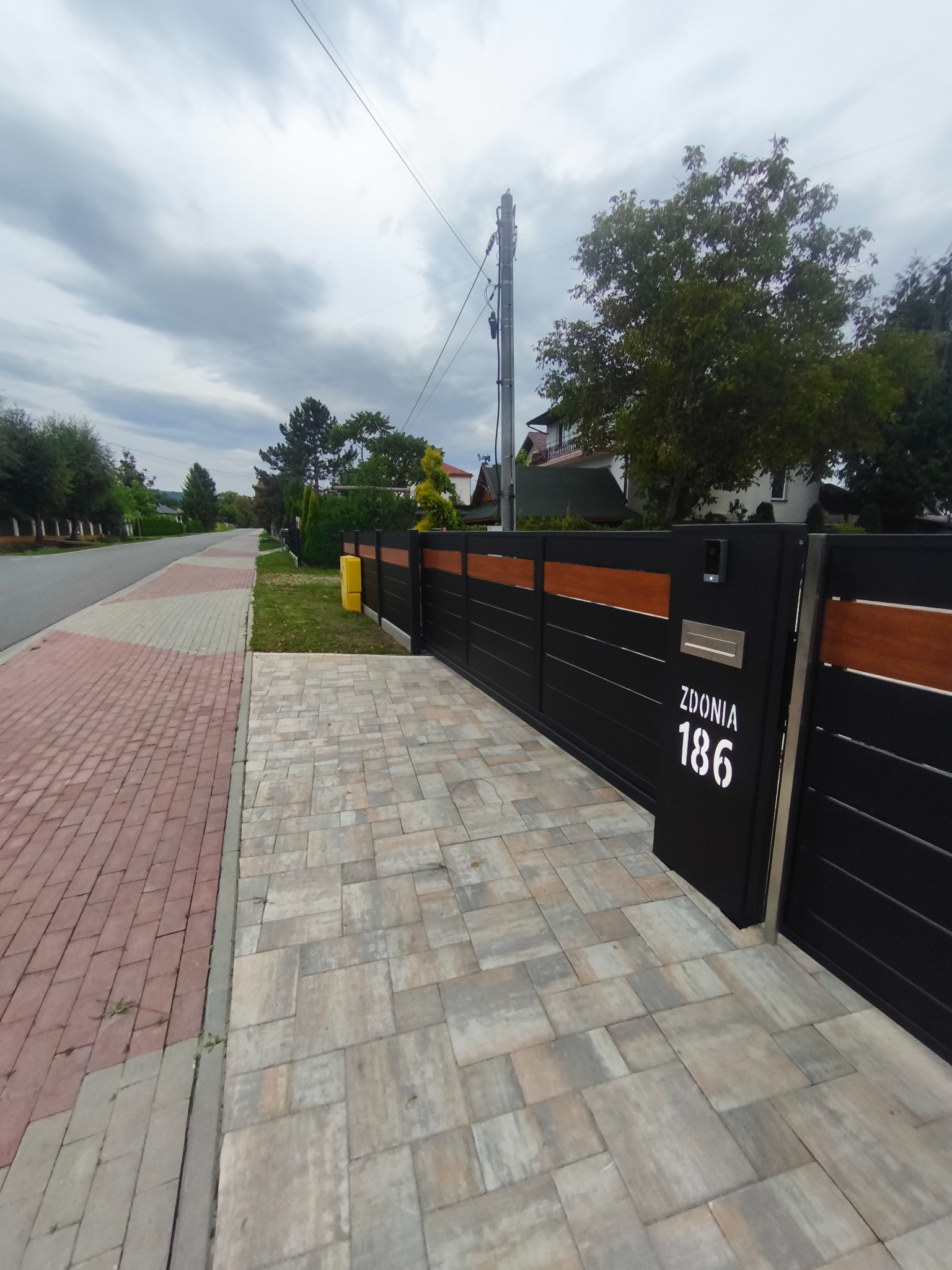 Nowoczesna brama wjazdowa z numerem domu 'Zdonią 186' w kolorze czarnym z drewnianymi akcentami. Brama przesuwna, widoczna z kostki brukowej.