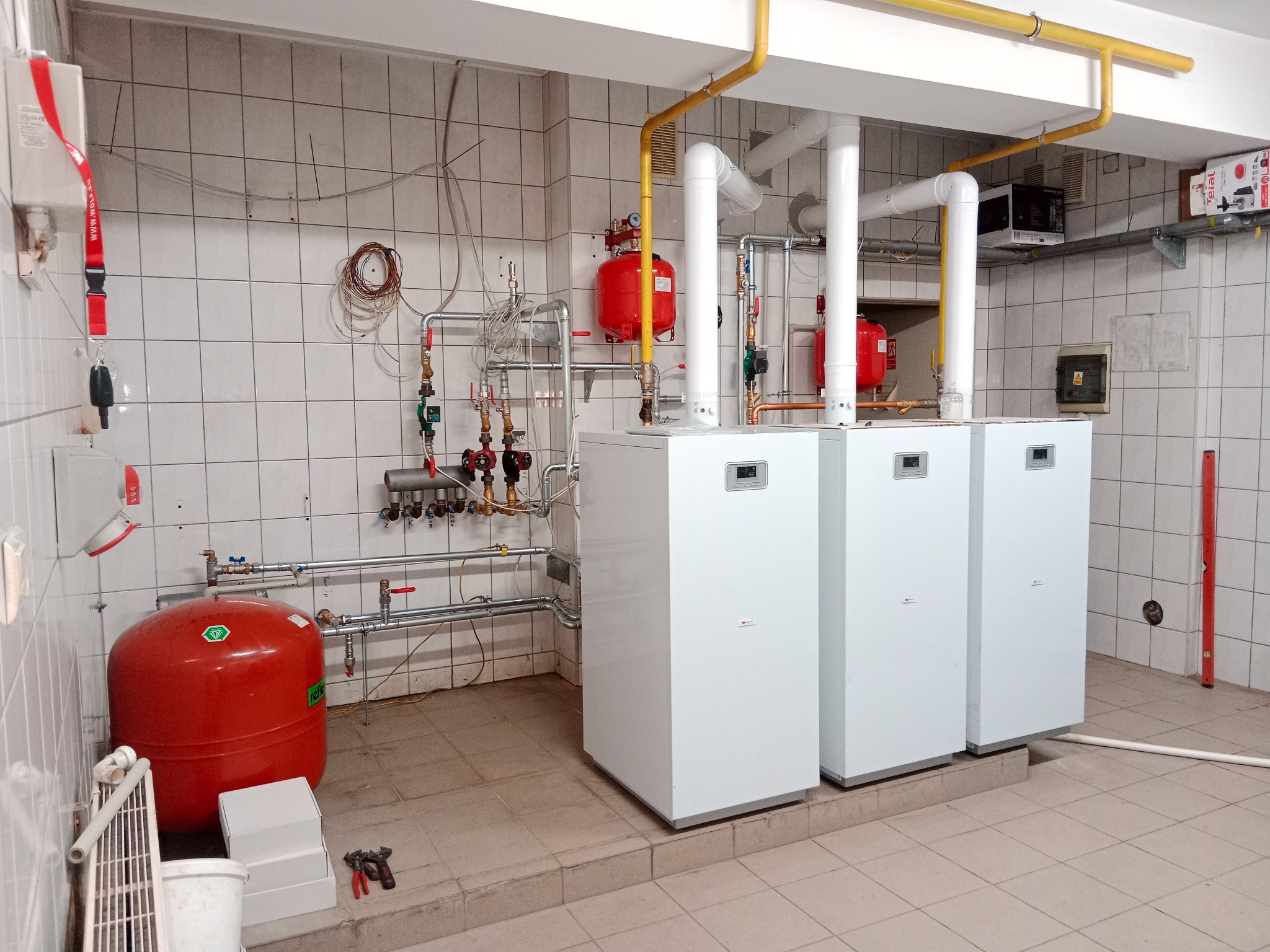 Trzy białe, nowoczesne piece gazowe zamontowane w pomieszczeniu z białymi kafelkami, widoczne rury gazowe w kolorze żółtym, czerwone zbiorniki wyrównawcze, instalacja wodna i poziomica oparta...