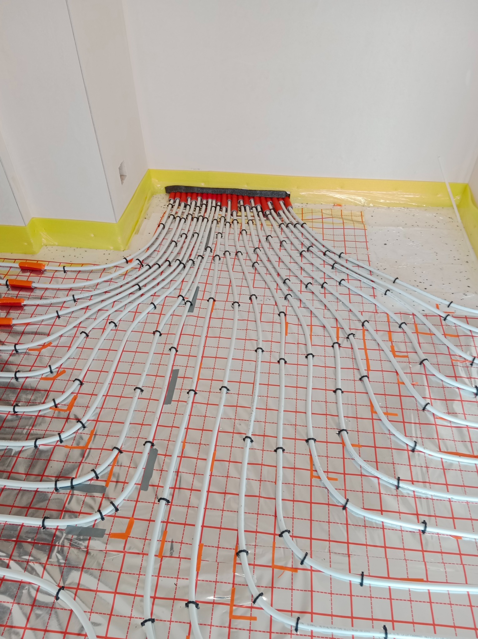 Białe rurki ogrzewania podłogowego rozłożone na folii z siatką, zabezpieczone czarnymi uchwytami, z czerwonym rozdzielaczem u góry, widoczne żółte folie zabezpieczające ściany.