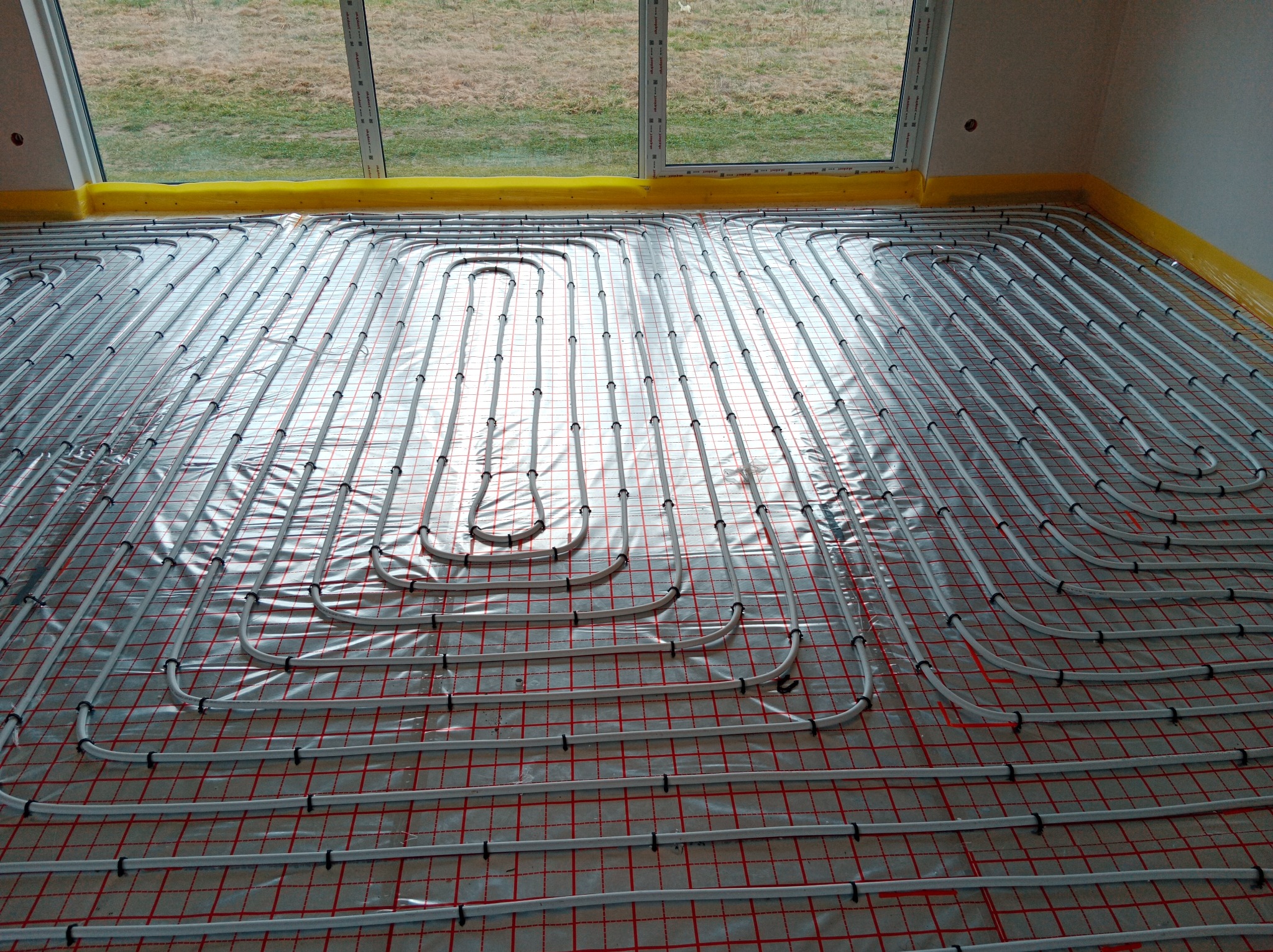 Białe rurki ogrzewania podłogowego ułożone w pętle na folii z czerwoną siatką, zabezpieczone żółtą taśmą przy ścianach i oknach, widok z góry.