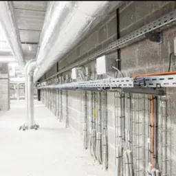 Korytarz techniczny z widocznymi instalacjami: rury wentylacyjne w srebrnej izolacji, metalowe korytka kablowe z przewodami, puszki elektryczne na ścianie z szarego betonu komórkowego.