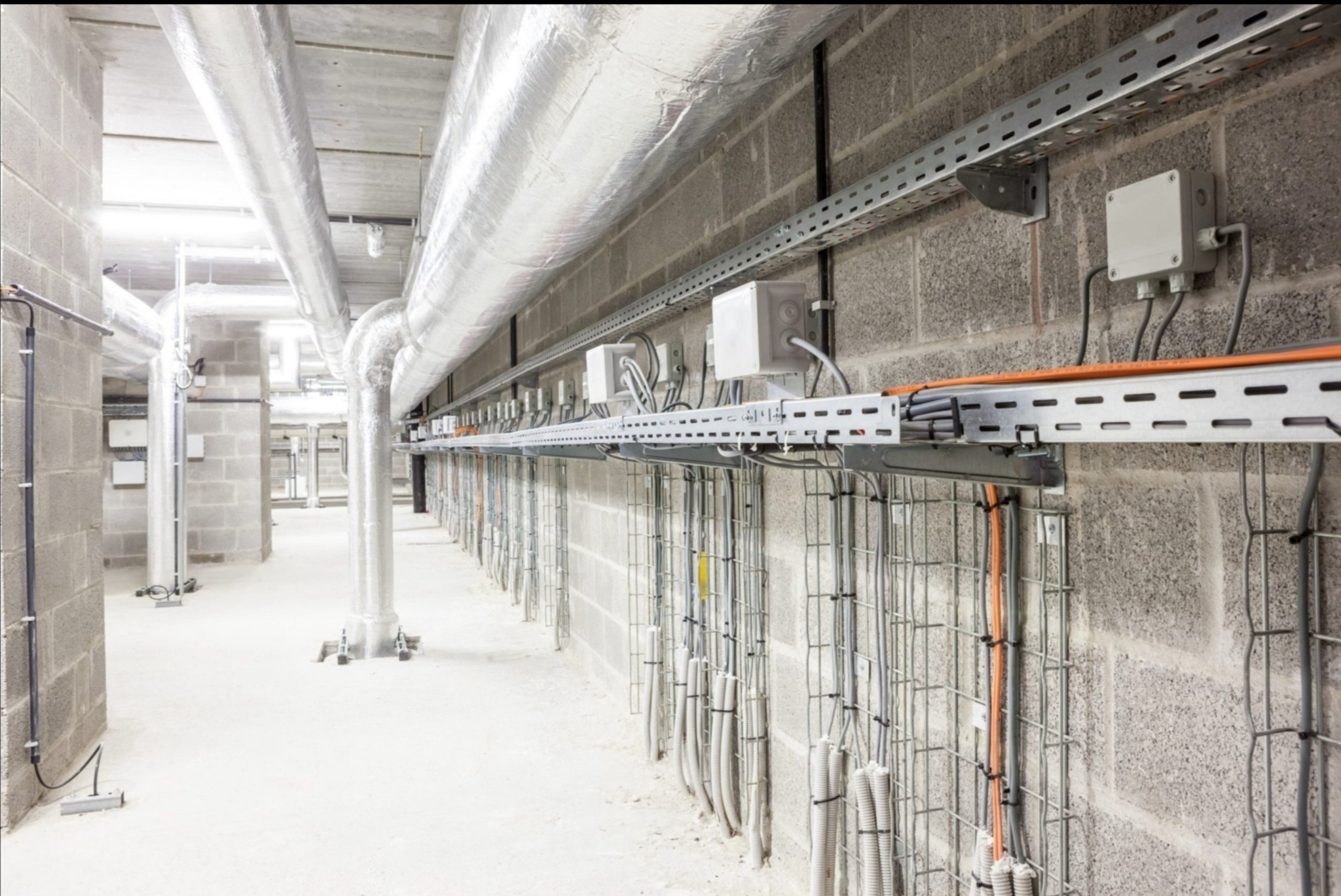 Korytarz techniczny z widocznymi instalacjami: rury wentylacyjne w srebrnej izolacji, metalowe korytka kablowe z przewodami, puszki elektryczne na ścianie z szarego betonu komórkowego.