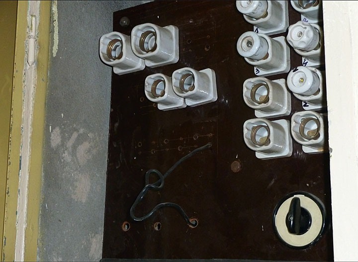 Tablica bezpiecznikowa z ceramicznymi gniazdami bezpieczników topikowych, widoczny kabel i przełącznik, zamontowana na brązowej płycie w pobliżu ściany.