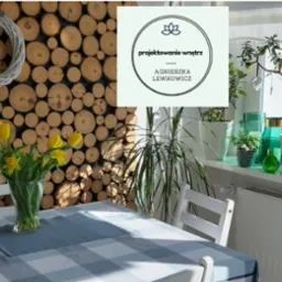 Jadalnia z drewnianą ścianą z okrągłych plastrów, bukietem żółtych tulipanów na stole w kratę, oraz roślinami doniczkowymi na parapecie okna, z logo firmy projektującej wnętrza.