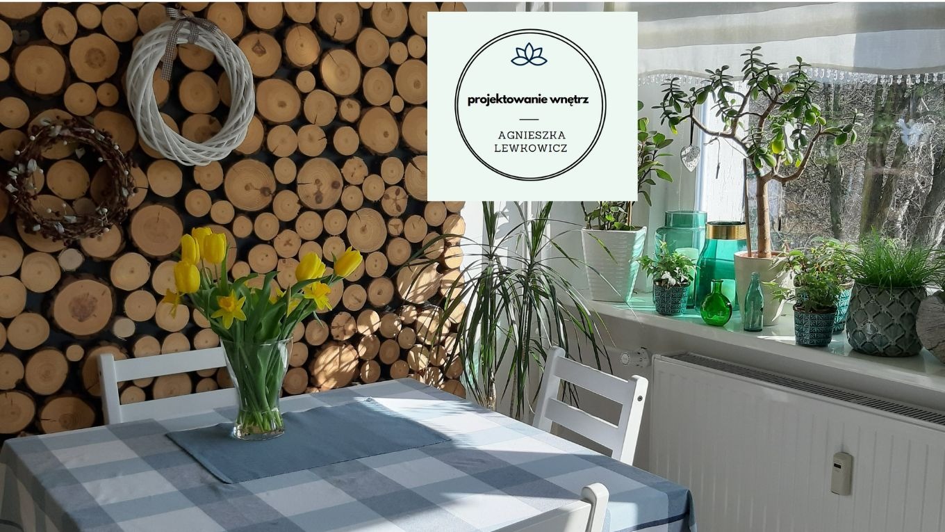 Jadalnia z drewnianą ścianą z okrągłych plastrów, bukietem żółtych tulipanów na stole w kratę, oraz roślinami doniczkowymi na parapecie okna, z logo firmy projektującej wnętrza.