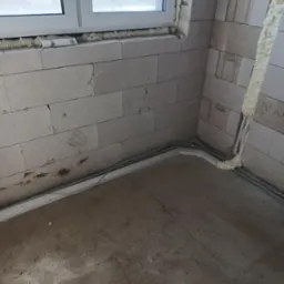 Surowy stan: ściany z betonu kom&oacute;rkowego z oknem, zabezpieczone pianką montażową. Widoczne szare kable instalacji elektrycznej w korytkach przy podłodze.