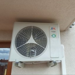 Arkadiusz Damięcki ADECO - Zamontowana jednostka zewnętrzna klimatyzacji CH z widokiem na miasto przez okno. Elewacja budynku o jasnej, chropowatej fakturze. Widoczne wsporniki mocujące.