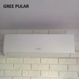 Biała klimatyzacja Gree Pular zamontowana na ścianie z białej cegły. Widoczne logo Gree i delikatna czerwona dioda sygnalizacyjna.