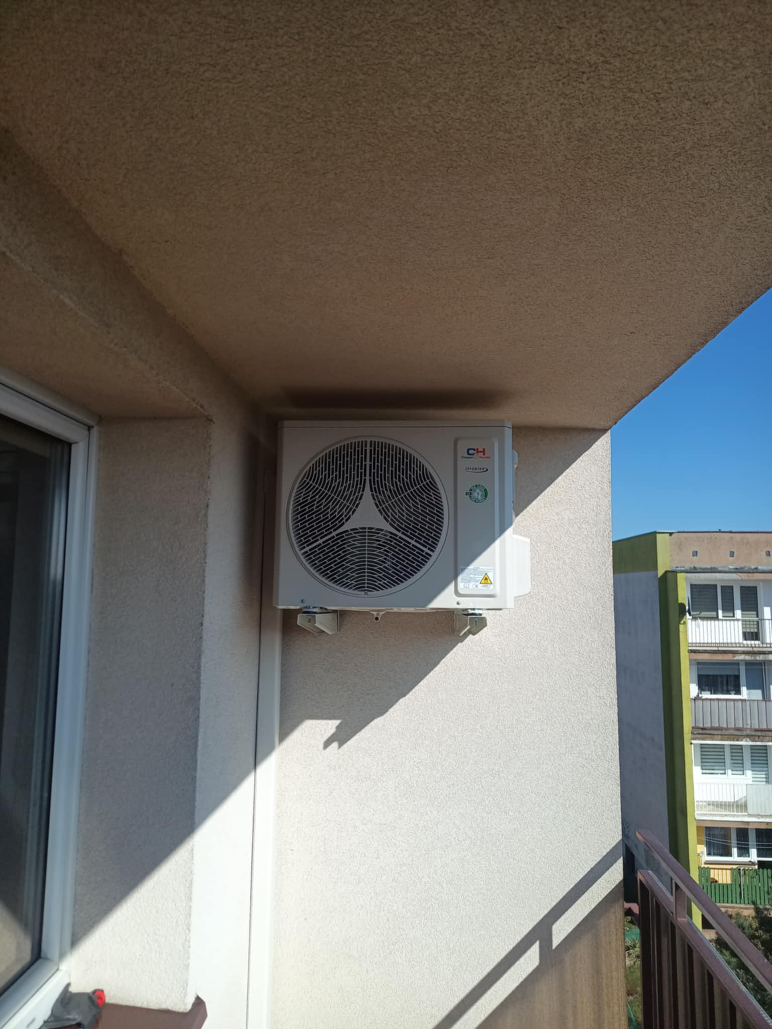 Zewnętrzna jednostka klimatyzatora CH Cooper&Hunter zamontowana na balkonie budynku mieszkalnego, widok z dołu pod kątem, fragment nieba w tle.