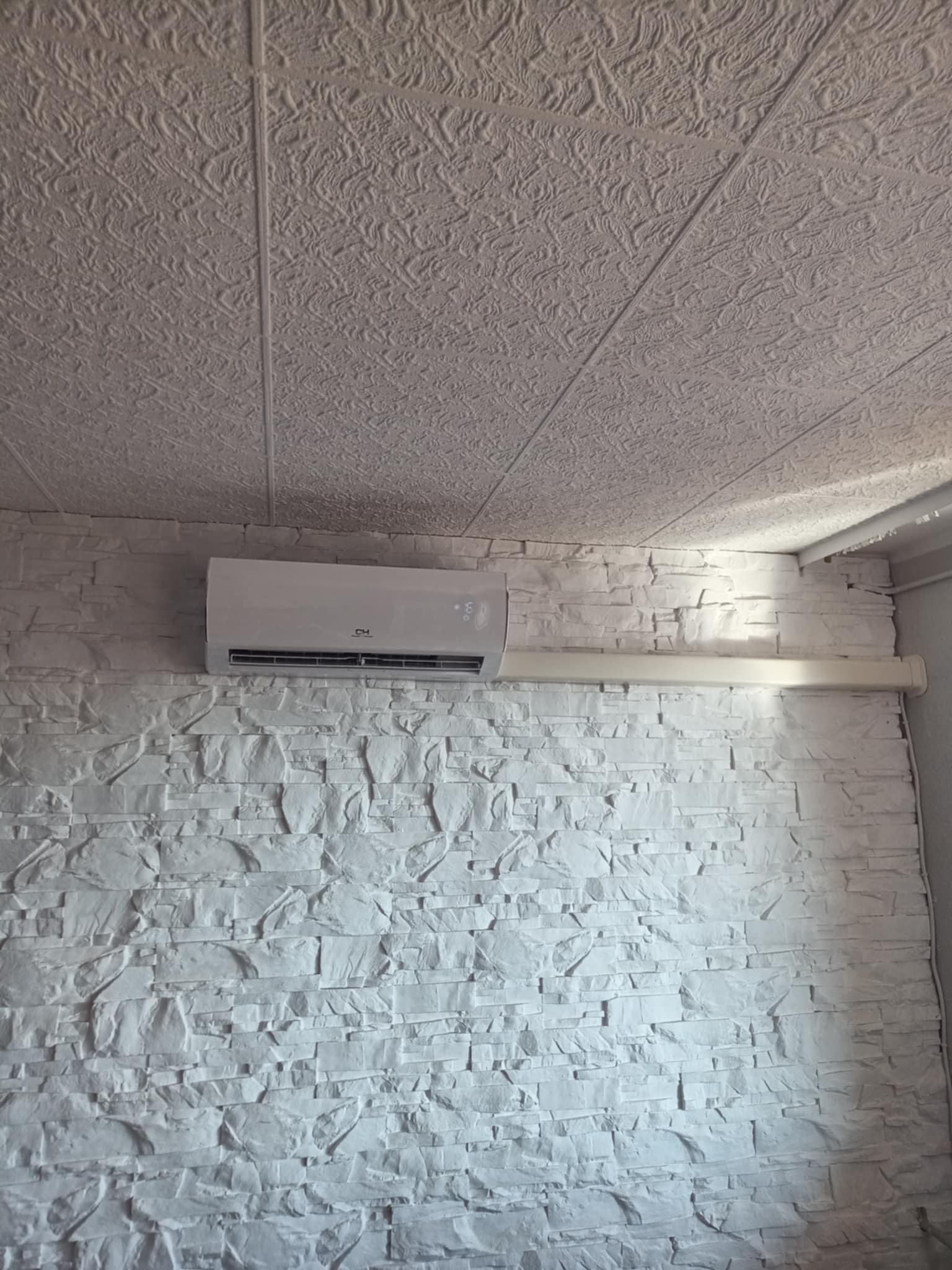 Biały klimatyzator marki CH zamontowany na ścianie z dekoracyjnej, białej cegły, pod sufitem z wzorzystą tapetą.