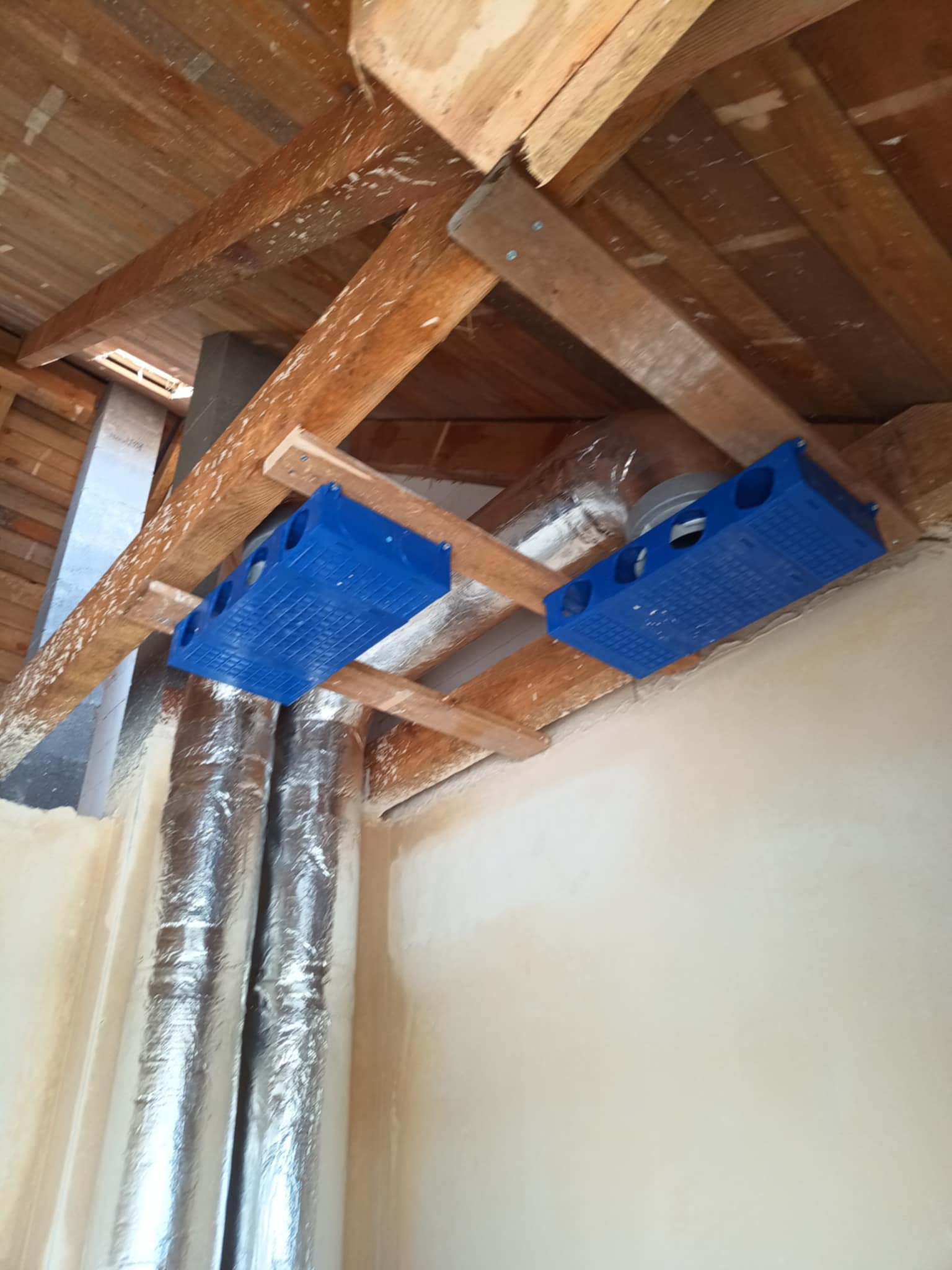 Niebieskie skrzynki rozdzielcze wentylacji zamontowane na drewnianej konstrukcji poddasza, z widocznymi rurami wentylacyjnymi w izolacji aluminiowej.