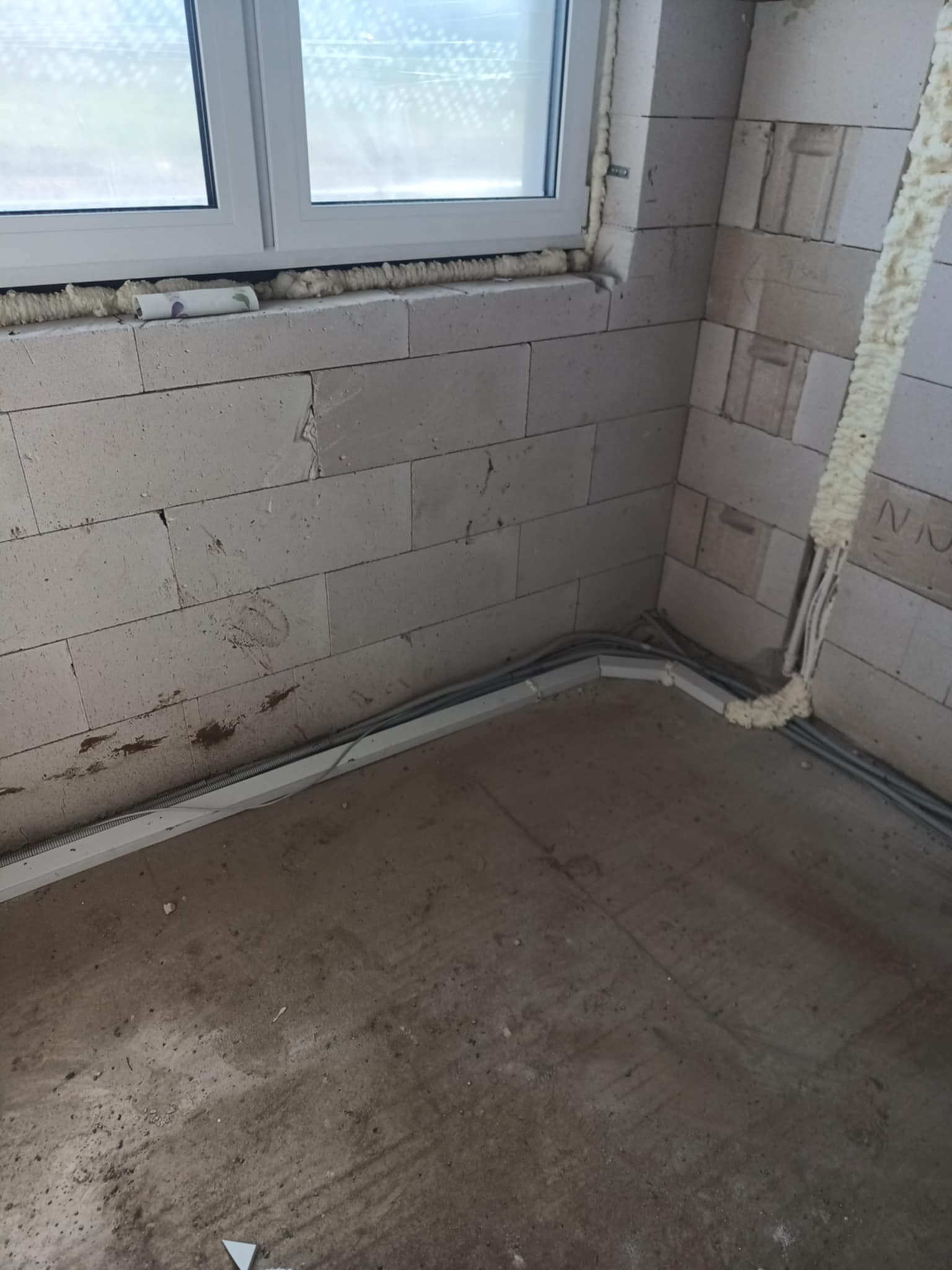 Surowy stan: ściany z betonu komórkowego z oknem, zabezpieczone pianką montażową. Widoczne szare kable instalacji elektrycznej w korytkach przy podłodze.