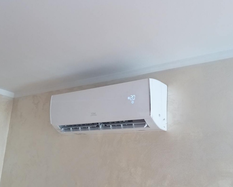 Biała klimatyzacja ścienna z wyświetlaczem temperatury 20 stopni, zamontowana w pomieszczeniu o jasnych ścianach i suficie.