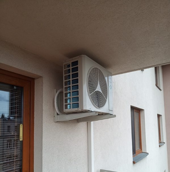 Biała jednostka zewnętrzna klimatyzacji zamontowana na ścianie budynku, widoczna z boku, z elementami instalacji. Obok okno z brązową ramą.