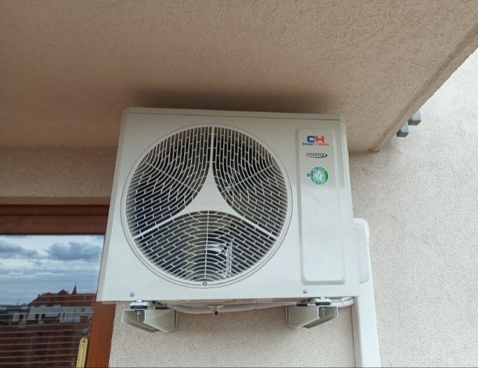 Zamontowana jednostka zewnętrzna klimatyzacji CH z widokiem na miasto przez okno. Elewacja budynku o jasnej, chropowatej fakturze. Widoczne wsporniki mocujące.