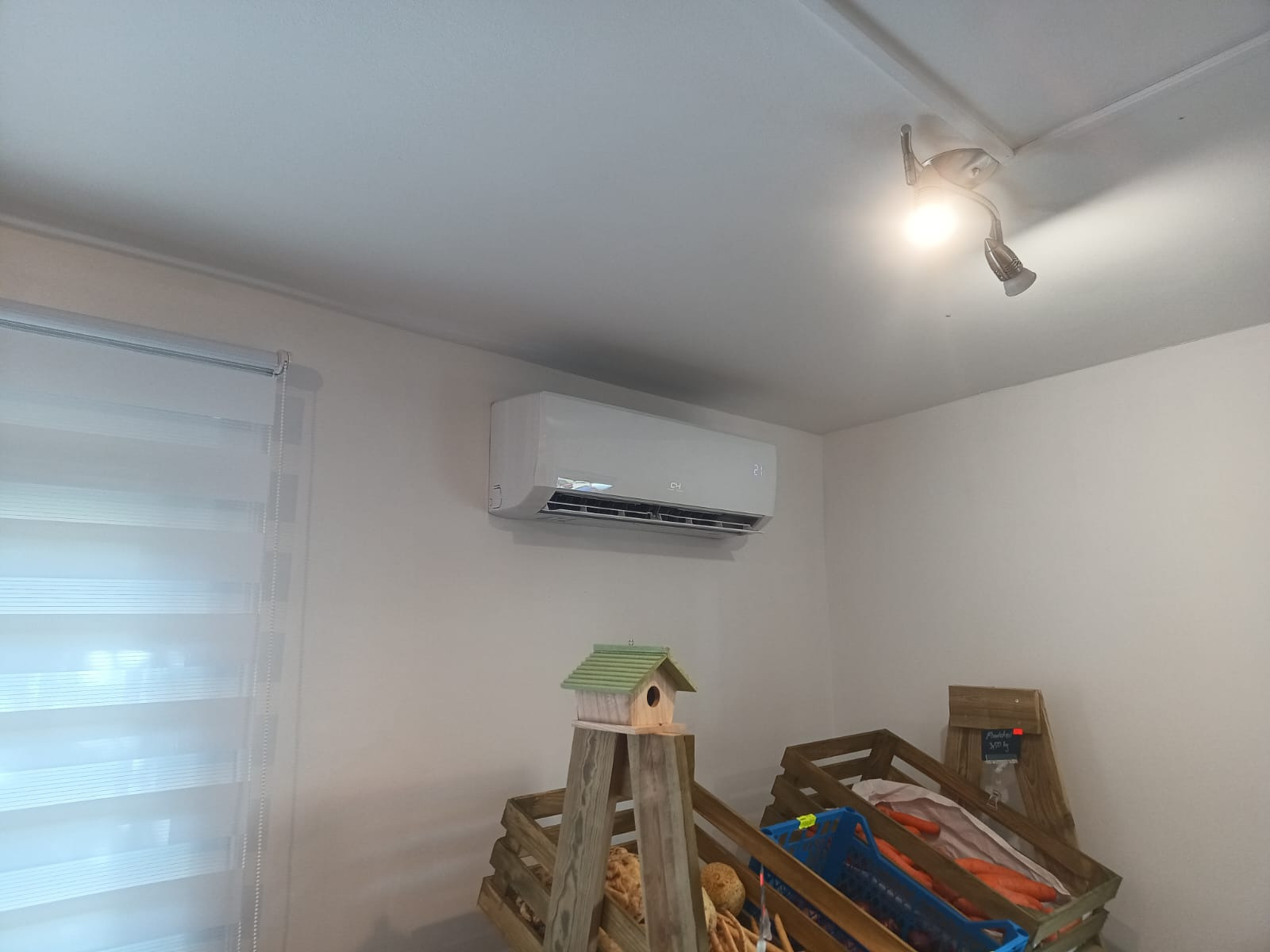 Biały klimatyzator ścienny w jasnym wnętrzu, widoczny na tle okna z roletami i dekoracyjnych skrzynek. Nowoczesny design, czyste linie, oświetlenie sufitowe.