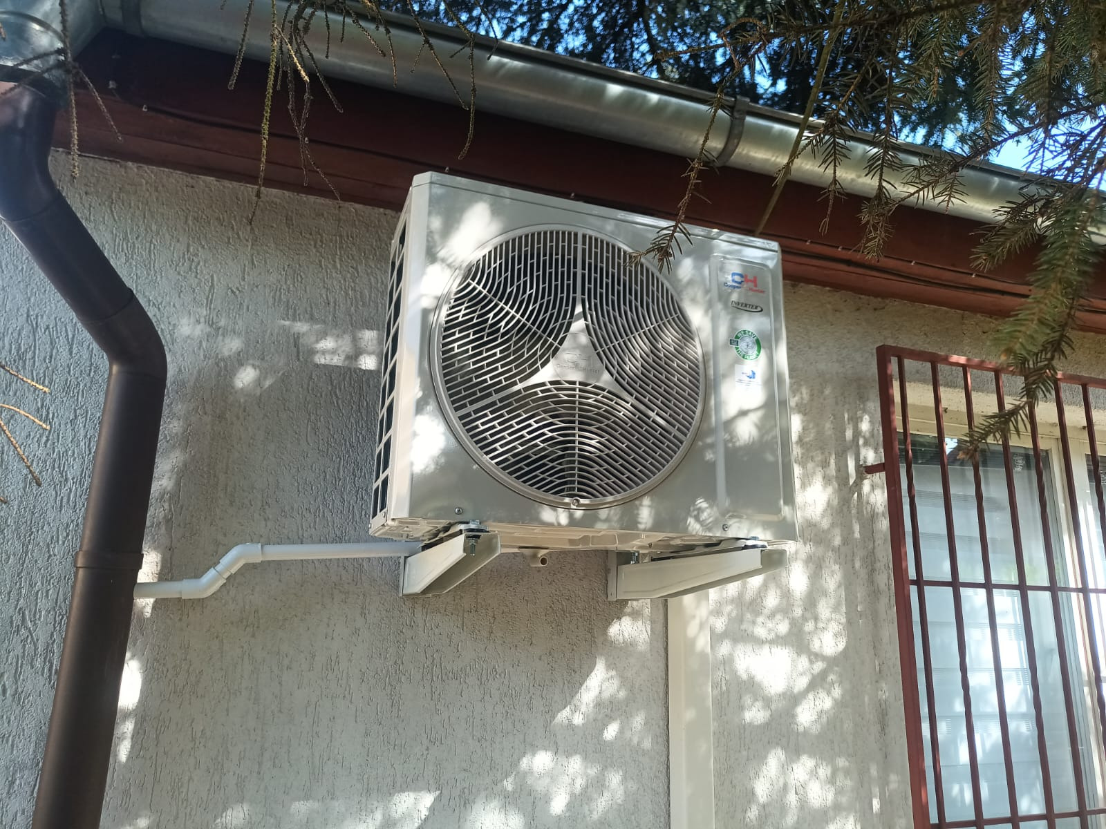 Zamontowana jednostka zewnętrzna klimatyzacji na elewacji budynku z brązową rynną, widoczne gałęzie drzewa i okno z kratą. Biała obudowa i wsporniki.