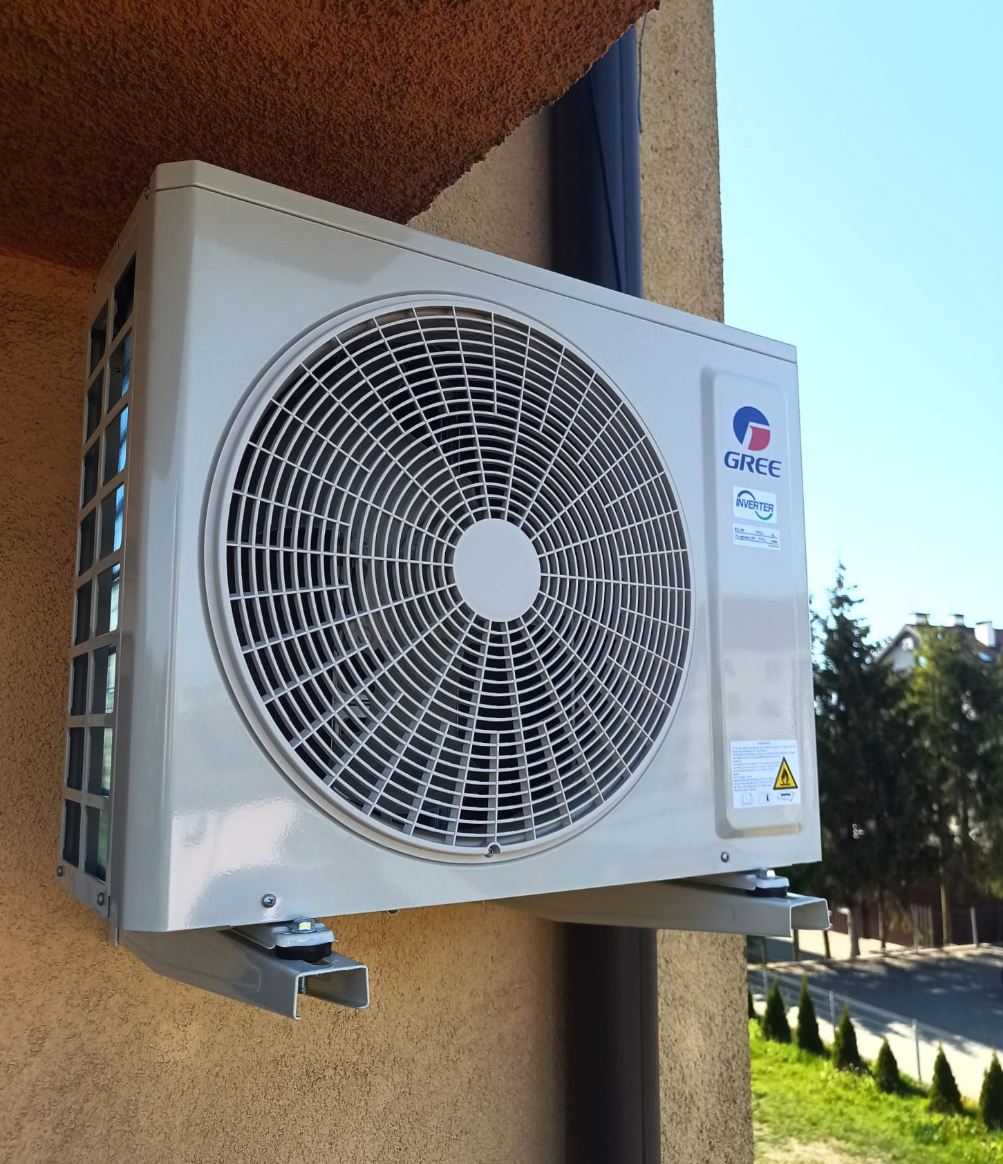 Zamontowana jednostka zewnętrzna klimatyzacji Gree na elewacji budynku. Widoczne mocowanie na metalowych wspornikach. W tle błękitne niebo i zieleń drzew.