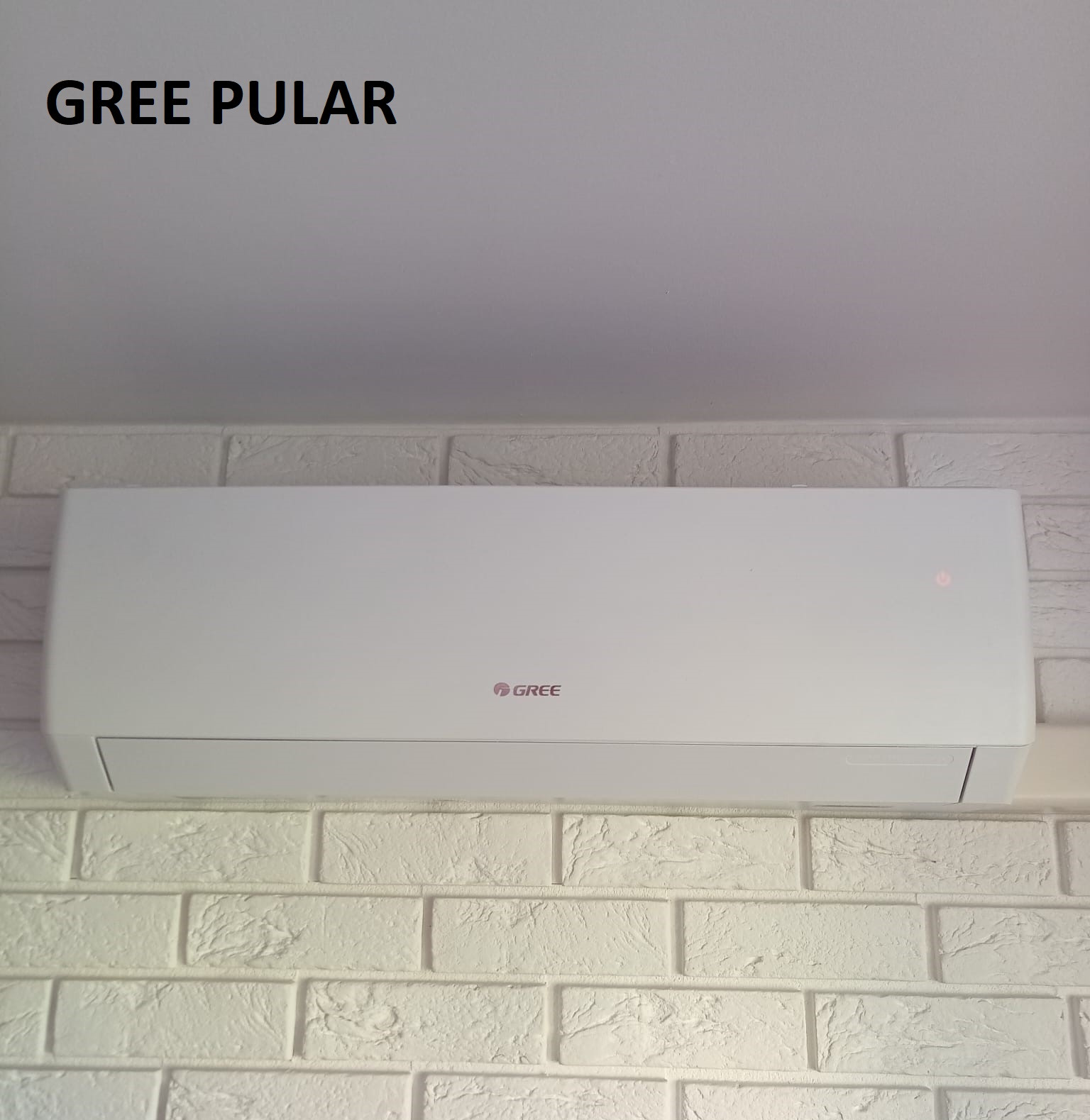 Biała klimatyzacja Gree Pular zamontowana na ścianie z białej cegły. Widoczne logo Gree i delikatna czerwona dioda sygnalizacyjna.