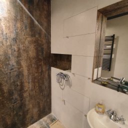 Eduard Ustymov - Łazienka w trakcie wykańczania: częściowo ułożone płytki w odcieniach beżu, ściana z brązową teksturą, widoczna umywalka i narzędzia glazurnicze.