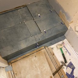 Eduard Ustymov - Układanie ciemnych płytek gresowych w narożniku pomieszczenia, widoczne krzyżyki dystansowe, młotek gumowy i opakowania po płytkach Ceramstic