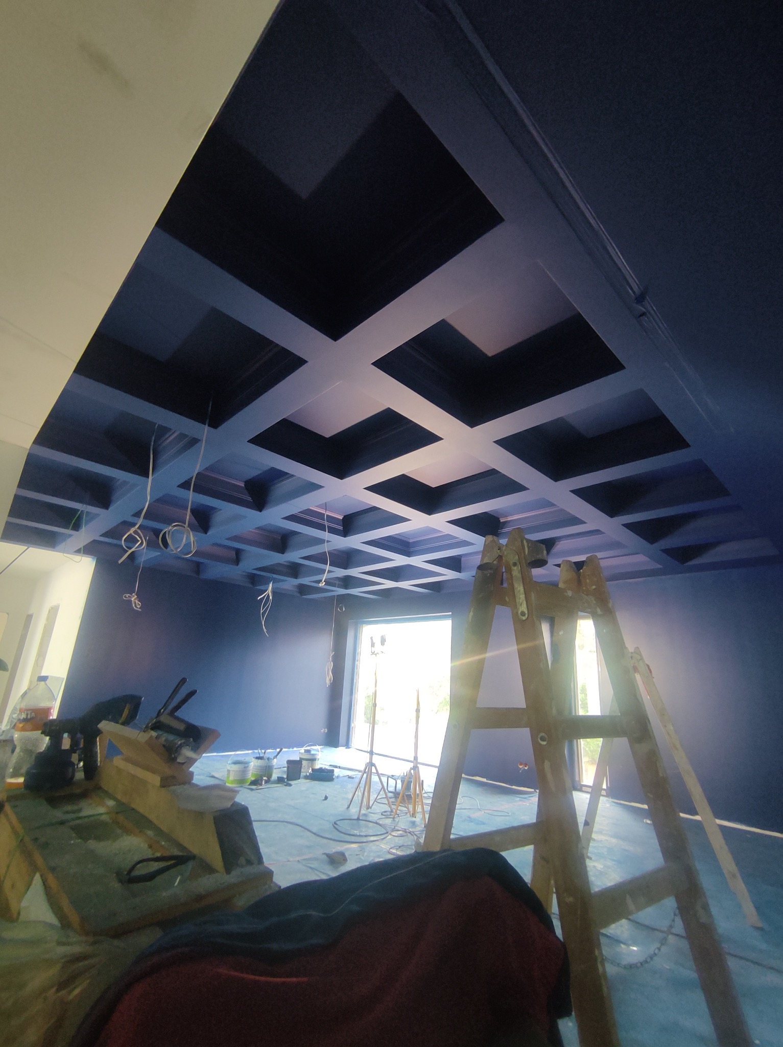 Niebieskie wnętrze w trakcie malowania z charakterystycznym kasetonowym sufitem, widoczna drabina malarska i narzędzia na podłodze, przewody zwisające z sufitu.