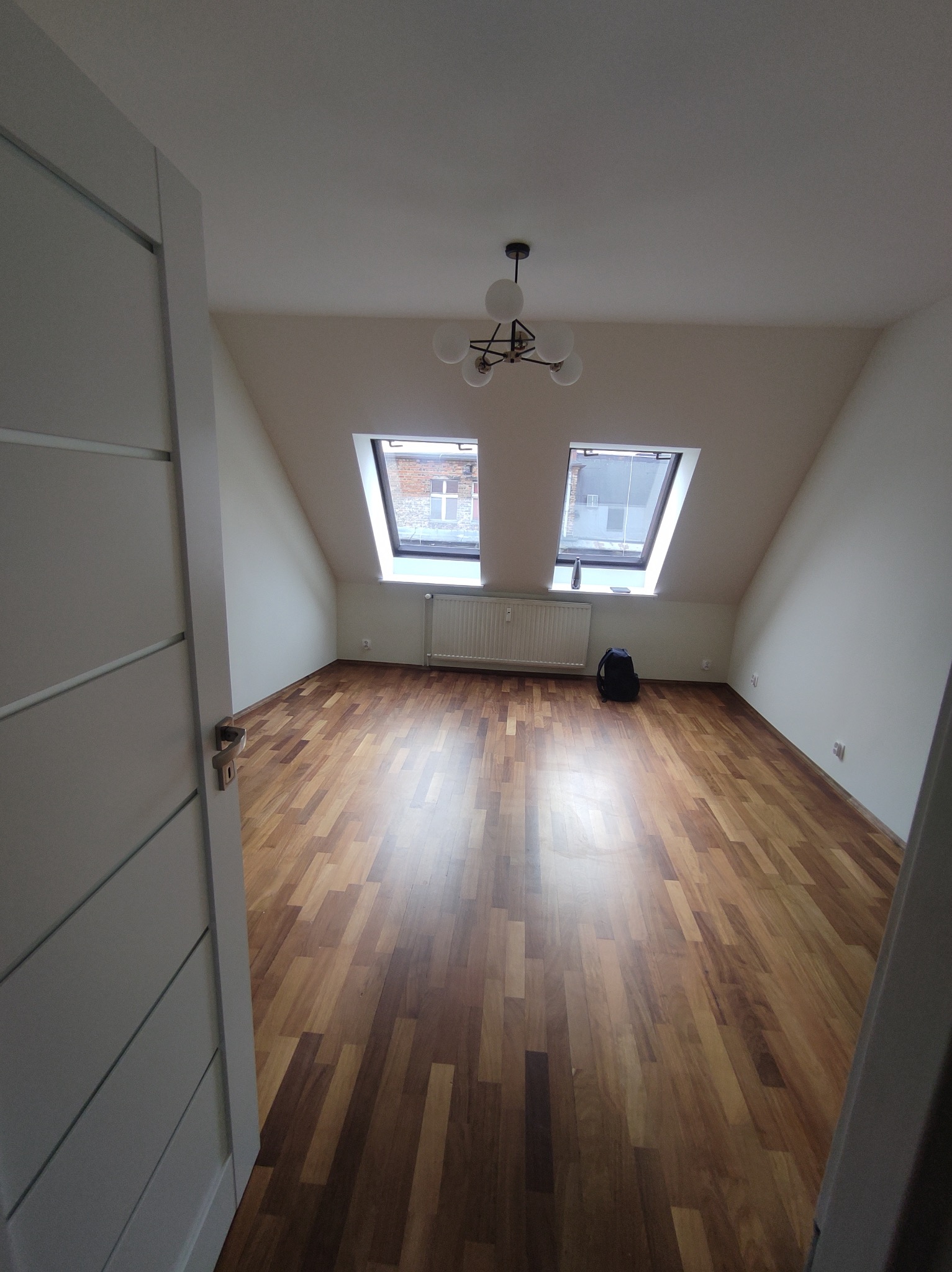 Poddasze po remoncie z drewnianą podłogą, dwoma oknami dachowymi i nowoczesną lampą sufitową, widok zza uchylonych drzwi.