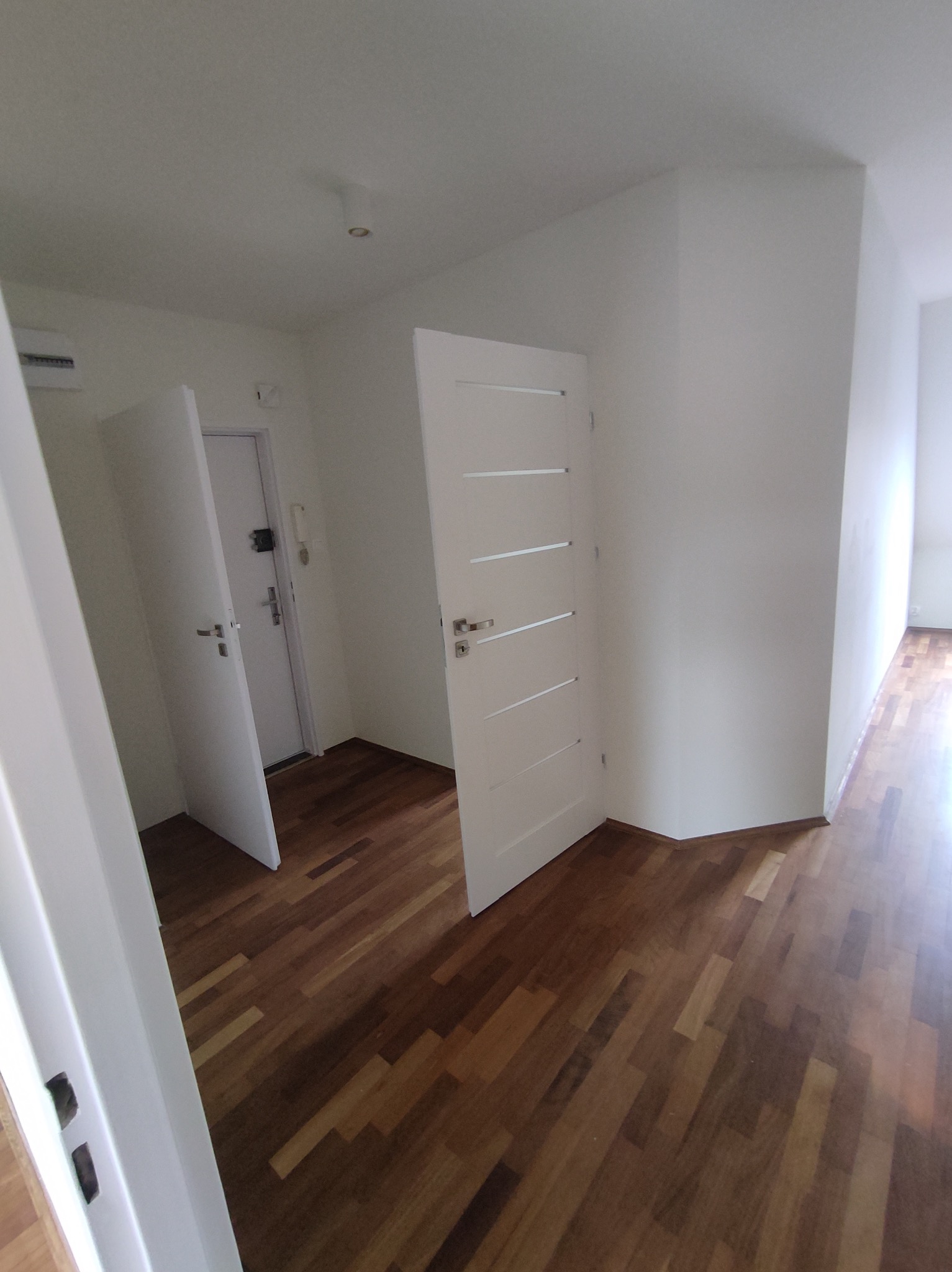 Wnętrze mieszkania po remoncie: widok na świeżo pomalowane białe ściany, drewnianą podłogę z parkietu ułożonego w jodełkę oraz dwie białe, częściowo otwarte drzwi.