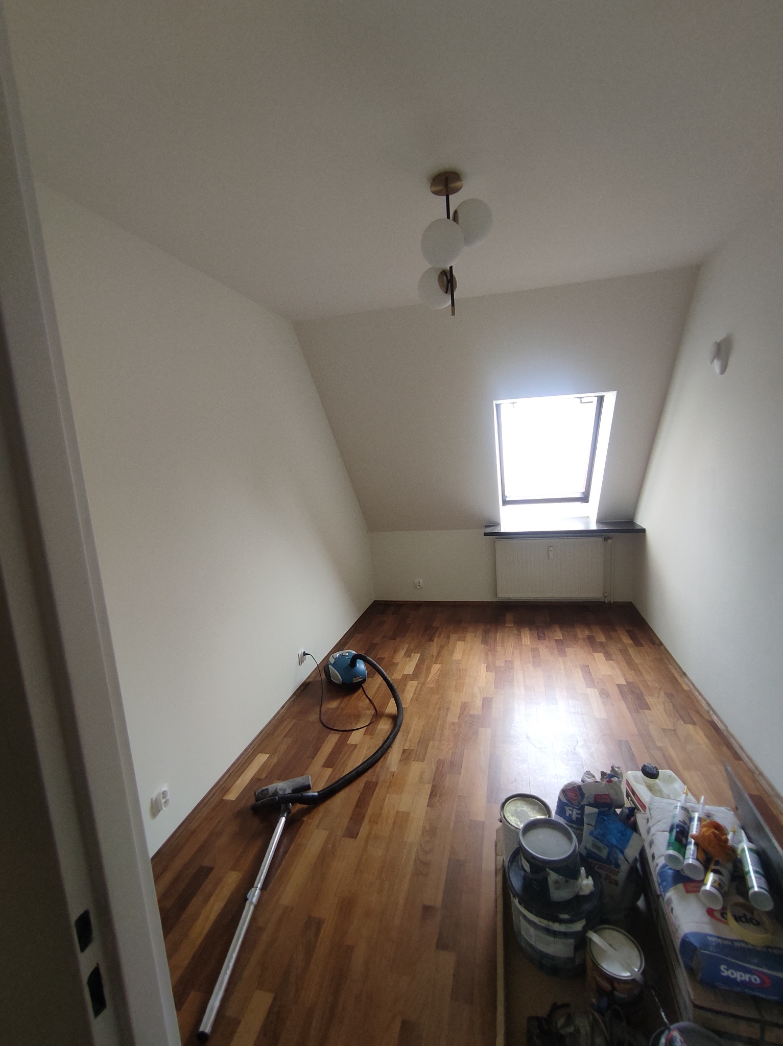 Pokój na poddaszu po malowaniu ścian na biało, z drewnianą podłogą, oknem dachowym, widocznym odkurzaczem i wiaderkami z farbą oraz innymi materiałami malarskimi.