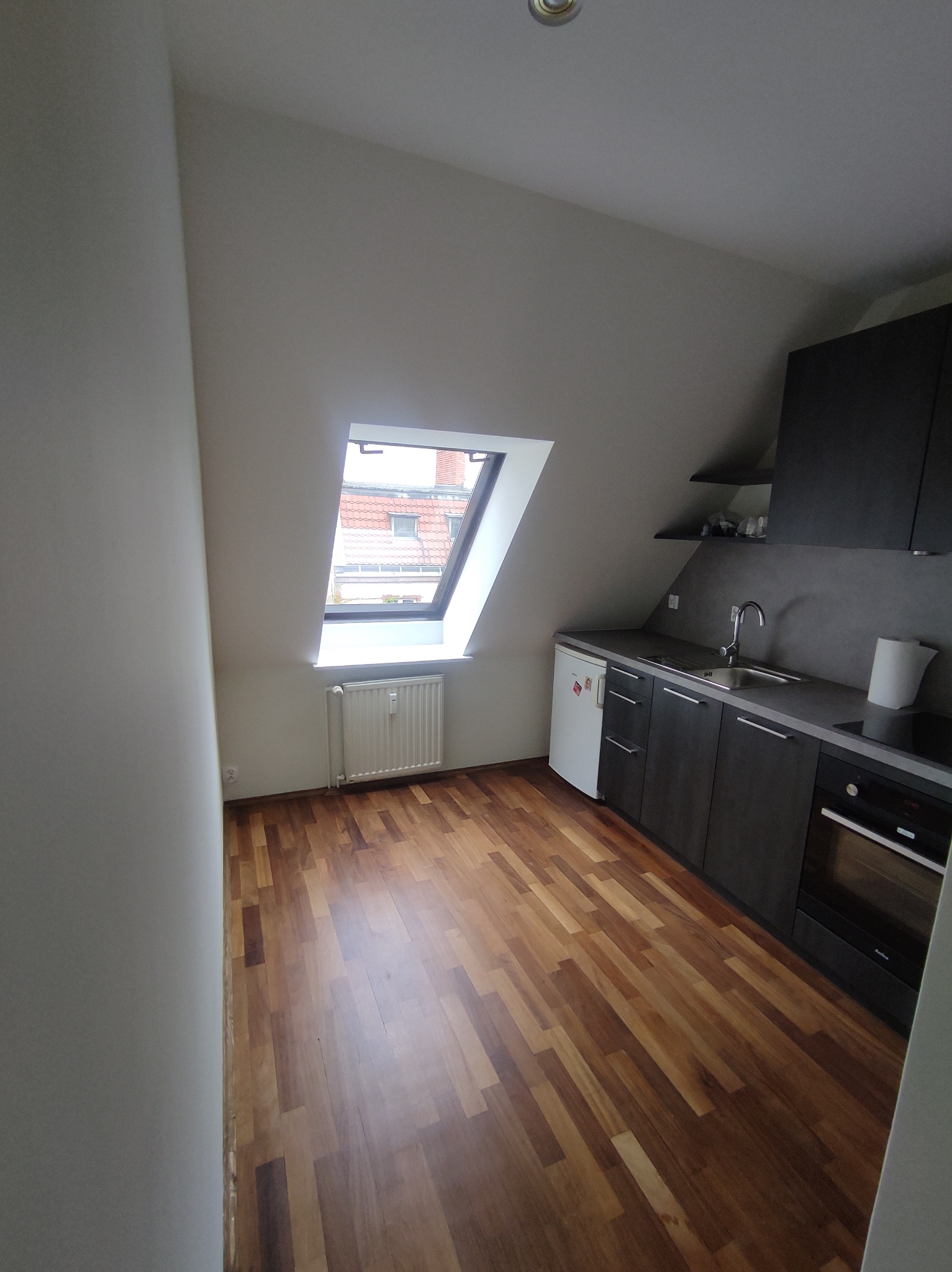 Nowoczesna kuchnia w skosie z drewnianą podłogą, czarne meble kuchenne, biała lodówka pod blatem, okno dachowe z widokiem na czerwony dach budynku.