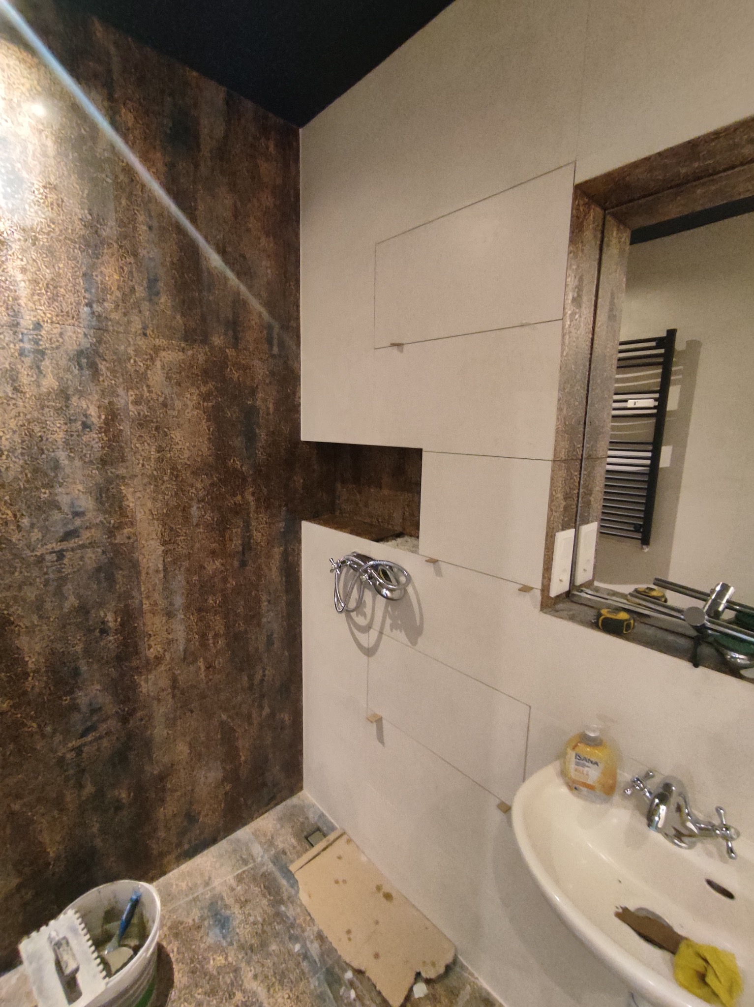 Łazienka w trakcie wykańczania: częściowo ułożone płytki w odcieniach beżu, ściana z brązową teksturą, widoczna umywalka i narzędzia glazurnicze.