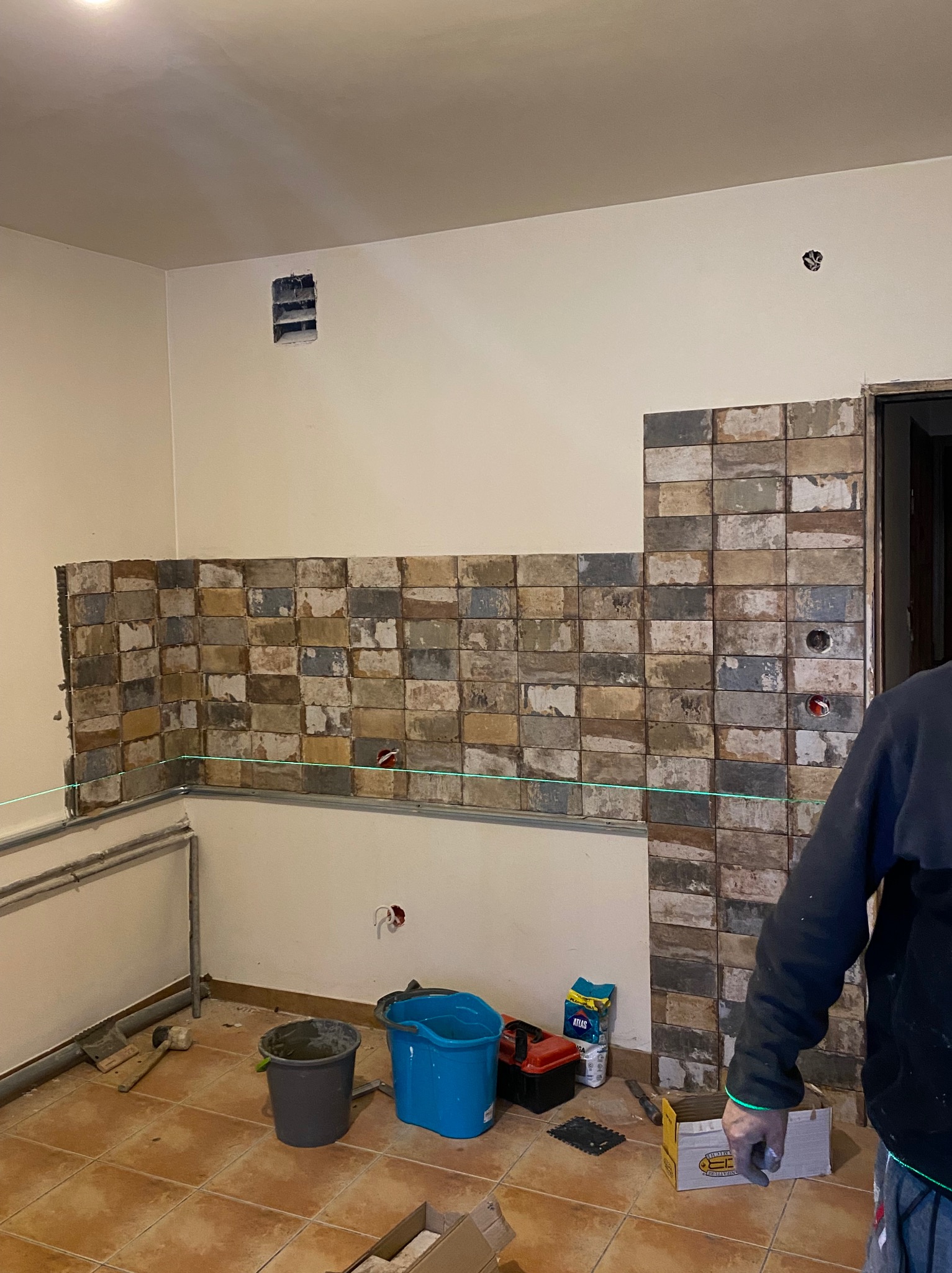 Układanie płytek imitujących cegłę na ścianie w kuchni, widoczny laserowy poziom, wiadra z zaprawą i narzędzia glazurnicze.