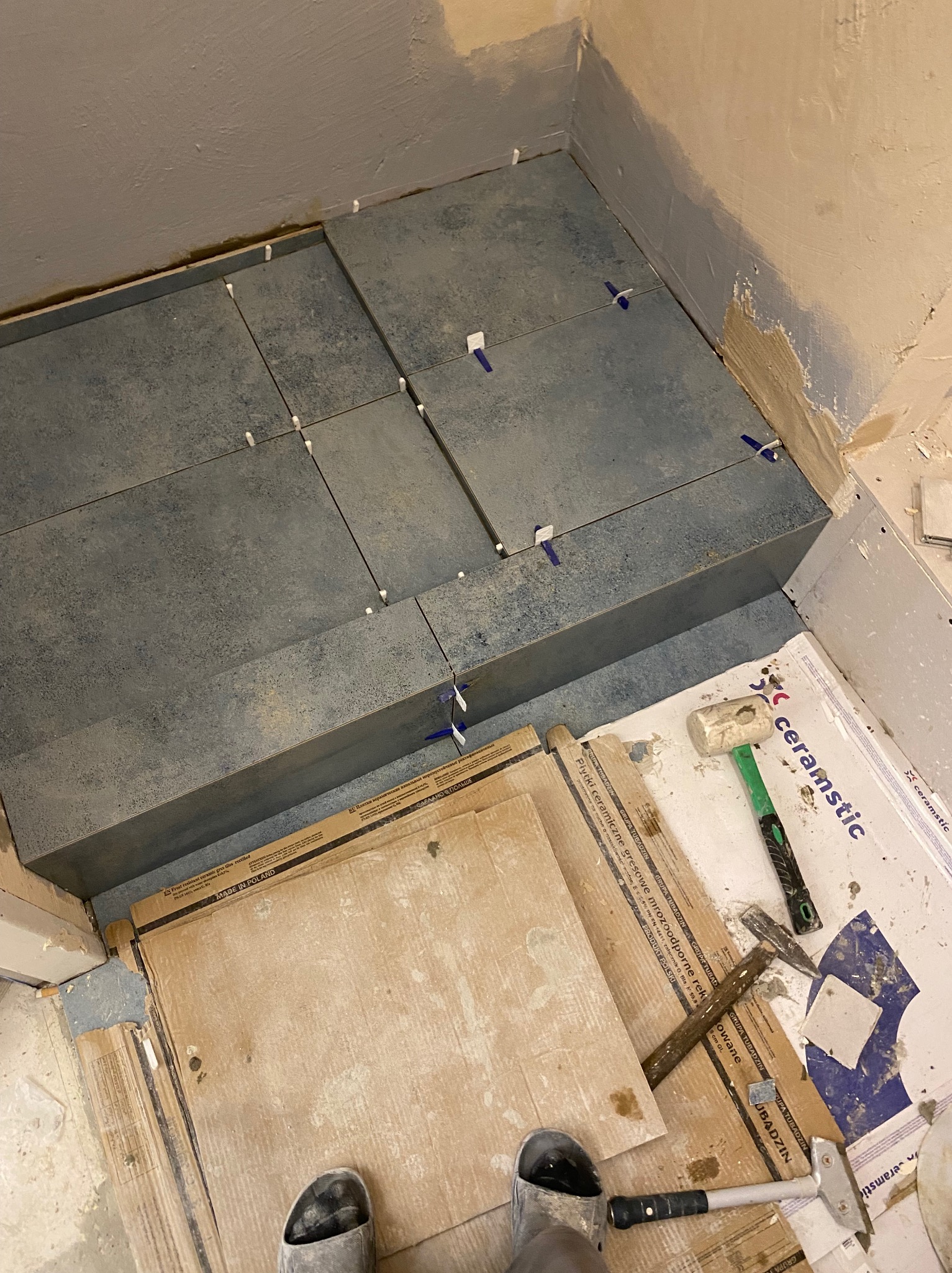 Układanie ciemnych płytek gresowych w narożniku pomieszczenia, widoczne krzyżyki dystansowe, młotek gumowy i opakowania po płytkach Ceramstic