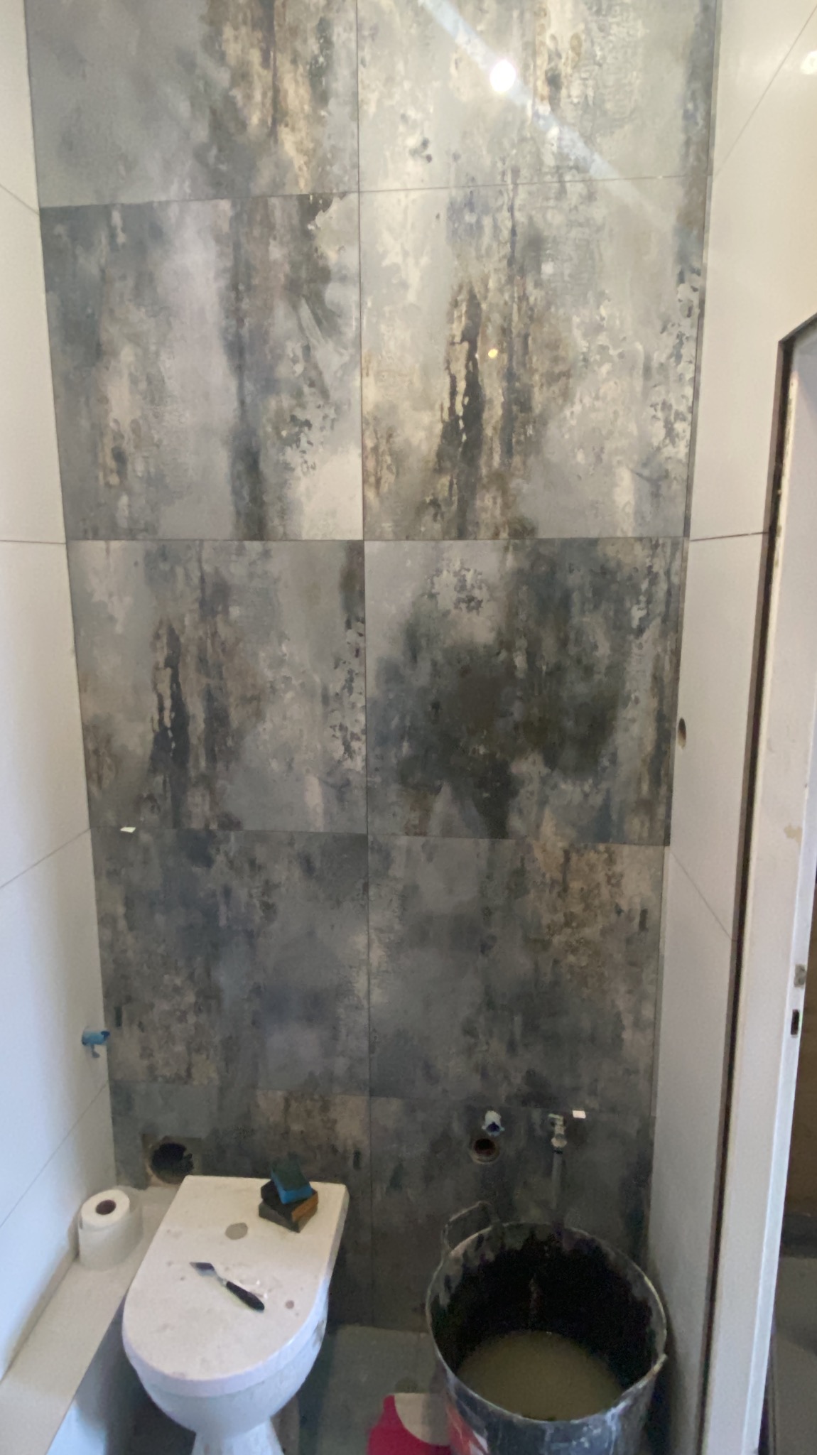 Łazienka w trakcie remontu: ściana wyłożona dużymi, szarymi płytkami imitującymi beton, widoczny fragment toalety z narzędziami i wiadro z zaprawą.